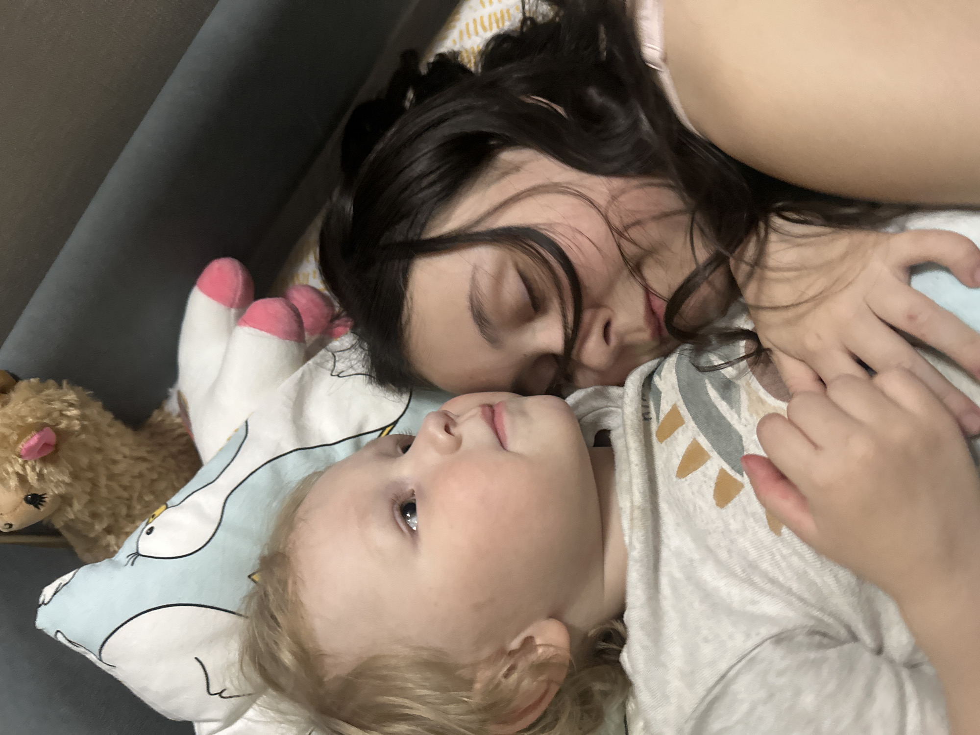 A child and an adult are lying on a bed. The child, who has blond hair and fair skin, is looking up at the adult. The adult, who has dark hair, has their eyes closed. The subjects are lying on a pillow with a cartoon rabbit design. A stuffed animal is to the left of the child. The location is not determined.