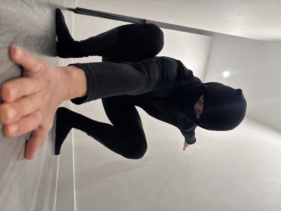A person in a black balaclava, black long-sleeved shirt, and black leggings poses in a room with white walls and a gray wood-look floor. The individual's left hand and right foot appear to be pressed against the floor and wall, creating a horizontal illusion. They are looking down at the camera with only their eyes visible. A door frame or trim appears in the background.