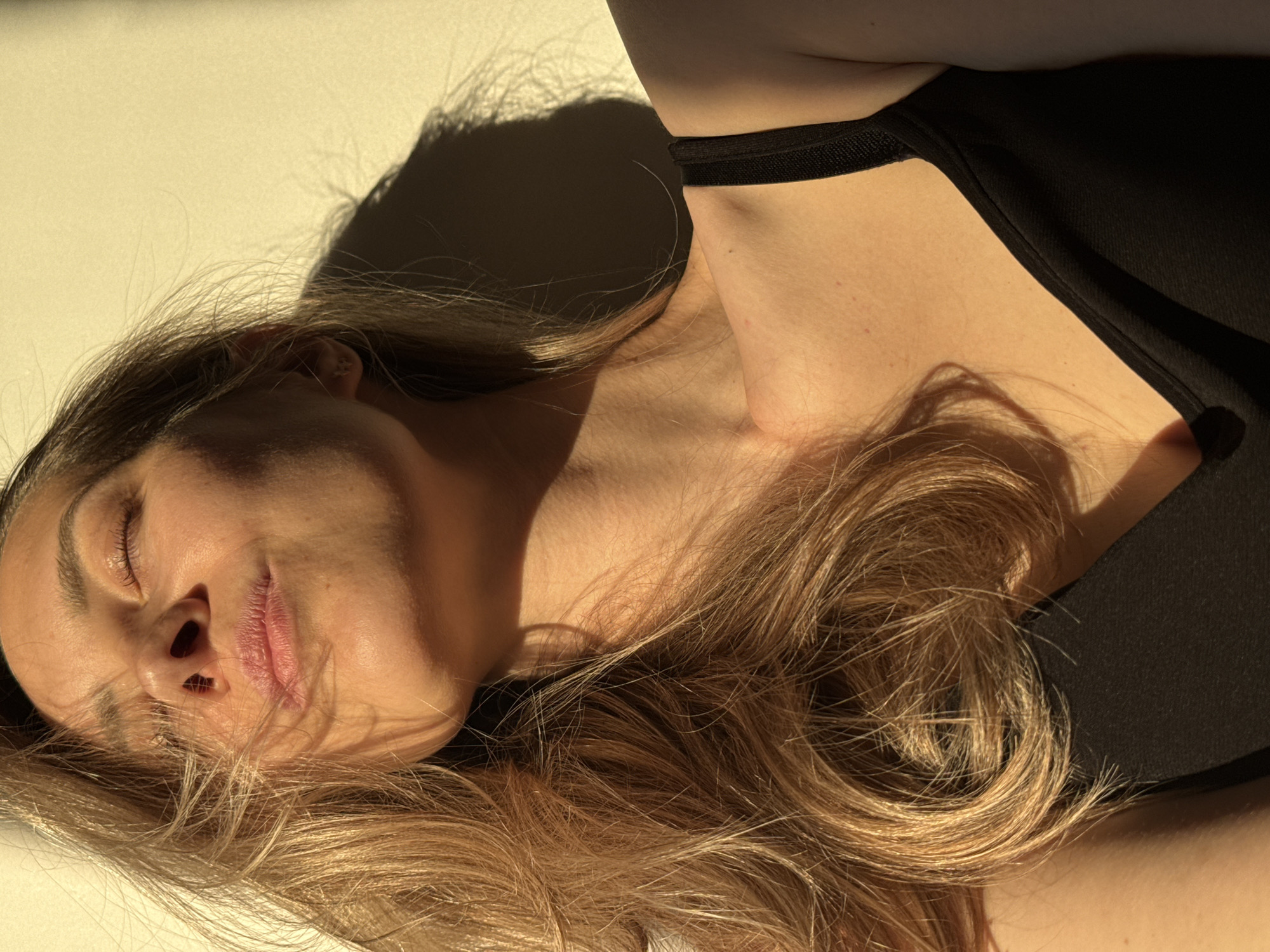 An individual, likely female, with long, wavy, light brown to blonde hair, is depicted lying down in a state of rest or sleep. Her eyes are closed, and her facial expression appears serene and relaxed. She is wearing a black garment, possibly a top or dress, with a thin strap visible over her shoulder. Her bare neck and upper chest are visible, illuminated by strong, warm light.
The scene is dominated by a clear, direct light source, casting prominent, sharp shadows from her head and hair onto a light, plain background, which appears to be a flat surface such as a floor or wall. These long shadows suggest the photograph was taken during either the early morning or late afternoon hours, under bright, sunny conditions. The intense sunlight creates a high-contrast effect, highlighting the textures of her skin and hair while deepening the shadows. Her hair is spread around her head and shoulders, with individual strands catching the light. No specific location cues are visible within the image itself, but the context provided places the scene in Chaikovsky, Russia. No visible text is present in the image.