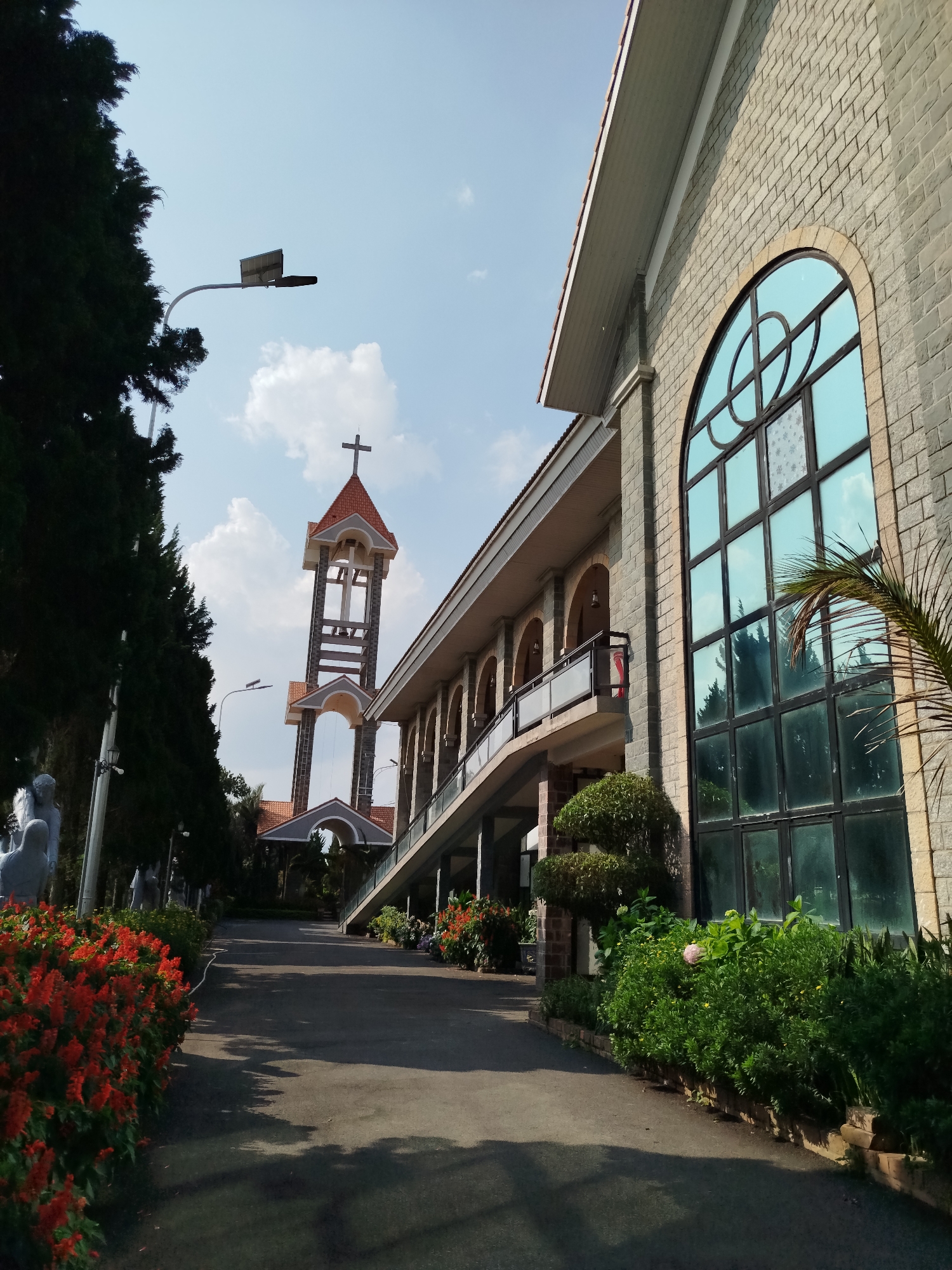 This image presents a tranquil outdoor view of a church complex in Diom A, Vietnam, bathed in bright sunlight under a clear, blue sky with scattered white clouds. The scene is devoid of people and captures architectural elements alongside vibrant natural landscaping.
To the right, a significant portion of a large church building, constructed with grey brick, is prominent. It features a striking tall, arched window with a circular design in its upper section and multiple rectangular panes below. This reflective window subtly mirrors the sky and surrounding trees. Below the building's roofline, a long balcony with arched supports and a modern glass railing extends along its side.
Further back and slightly to the left, a tall, modern bell tower stands out. It possesses an open, multi-tiered design, capped with a red-tiled roof and a visible Christian cross. Its base incorporates arched openings.
A paved pathway, likely asphalt, gently curves towards the bell tower, bordered by lush greenery. In the immediate foreground on the left, a dense bed of bright red flowers, possibly Salvia, adds a vivid splash of color. Tall, dark, conifer-like trees line this side of the path, partially obscuring several white or light-colored statues that appear to be religious figures. Various green shrubs and plants are abundant throughout the grounds, including a palm frond visible on the far right. A streetlight, equipped with what appears to be a small solar panel, is visible against the sky.
The scene evokes a sense of peacefulness and reverence, characteristic of a religious sanctuary during the daytime. No specific activity or event is taking place, and no visible text is present.