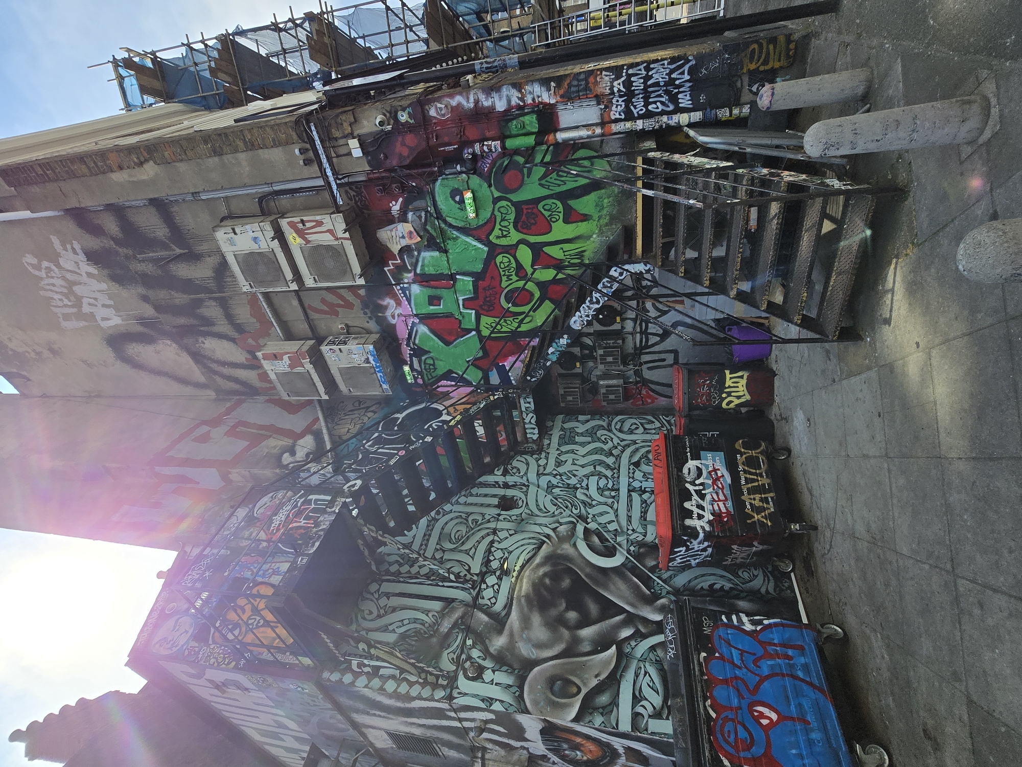 The image captures a narrow alleyway in London, characterized by vibrant graffiti art covering the walls and surfaces. A metal staircase ascends the right side, leading to upper levels, and a series of air conditioning units are attached to the building's facade. The ground level includes several wheeled containers, also decorated with graffiti. Scaffolding is visible at the top of the frame. The overall impression is one of urban grit and artistic expression.