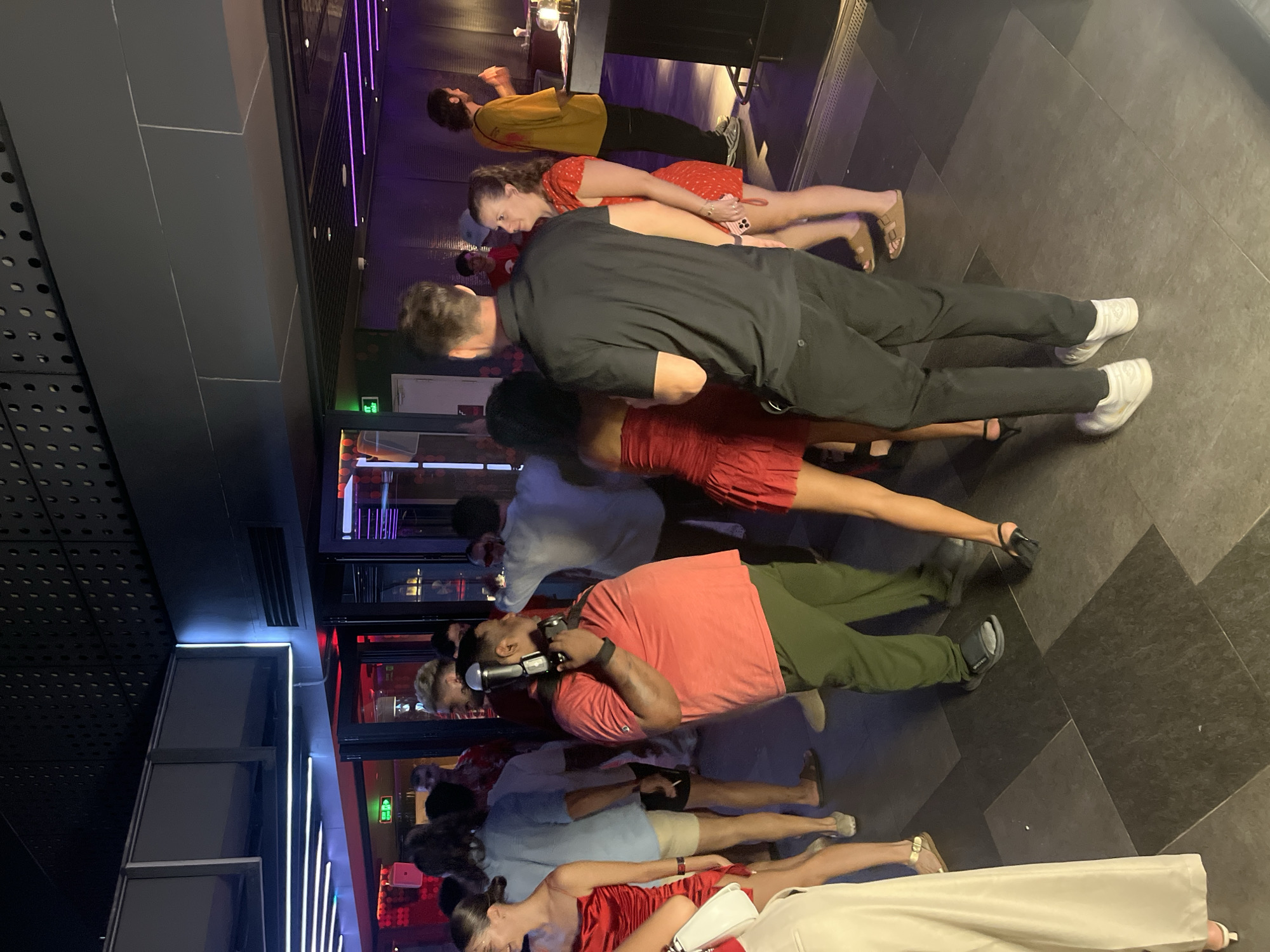 A group of individuals are gathered indoors, possibly at a club or event venue, in Da Nang, Vietnam. The scene is dimly lit with purple and white neon lighting accentuating the modern architectural design of the space. In the foreground, a man wearing a coral-colored t-shirt and green pants is holding a camera with a flash attachment, appearing to be taking a photograph. He is positioned to the left of a man in a black polo shirt and black pants. Several women are visible, many wearing red dresses. One woman in a red dress stands to the right of the man in the black polo shirt, while another woman in a similar dress is partially visible on the far left. Other individuals are scattered throughout the background, some engaged in conversation. The floor is tiled with dark, large square tiles. The overall atmosphere suggests a social gathering or entertainment event.
