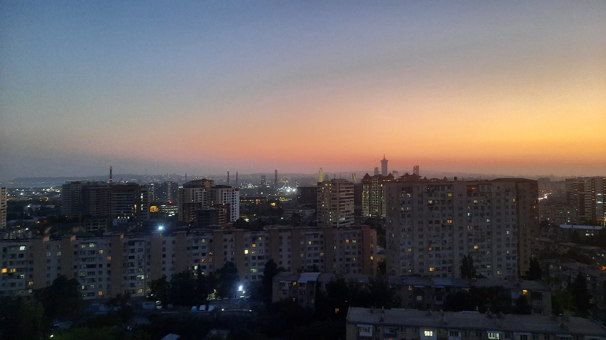 An elevated panoramic view captures a city skyline at dusk. The sky transitions from a deep blue at the top to a warm orange and yellow gradient near the horizon, indicating the setting sun. In the foreground, several multi-story apartment buildings are visible, their windows illuminated with a warm, yellow light. Some buildings appear older with more uniform structures, while others are modern high-rises. In the mid-ground and background, more buildings and infrastructure are discernible, including industrial elements such as smokestacks or cooling towers, some emitting light. The overall scene depicts an urban environment transitioning from daylight to evening, with the city lights beginning to twinkle against the fading natural light. The image is taken from a high vantage point, offering a wide perspective of the cityscape.