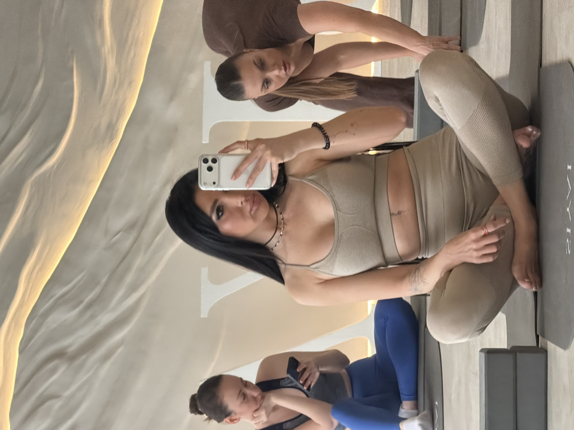 This image depicts three women within a modern yoga studio identified as Jayavic, located at Novy Arbat, 21, Moscow, Russia.
In the foreground, a woman with long dark hair is seated barefoot in a cross-legged position on a dark gray yoga mat, wearing a beige sports bra and matching leggings. She holds a white smartphone with her left hand, taking a mirror selfie. She wears a black choker, a beaded necklace, a black coiled bracelet on her left wrist, and has visible tattoos on her left forearm.
To her left, a second woman with dark hair pulled into a bun is seated cross-legged on a separate yoga mat, dressed in a dark blue sports bra and leggings with white socks. She is looking down at a black smartphone held in her hands. Two gray yoga blocks are stacked on the floor adjacent to the foreground subject's mat.
To the right, a third woman with light brown hair in a ponytail is partially visible, bent forward in an exercise pose, wearing a brown ribbed top and matching pants.
The studio features light-colored flooring and walls. Large, stylized white letters, partially visible on the back wall, spell out "JAYAVIC." The ceiling displays undulating white panels with integrated warm strip lighting, creating an illuminated, wave-like pattern. Each participant occupies an individual yoga mat; the mat in the foreground bears the inverted "JAYAVIC" logo.