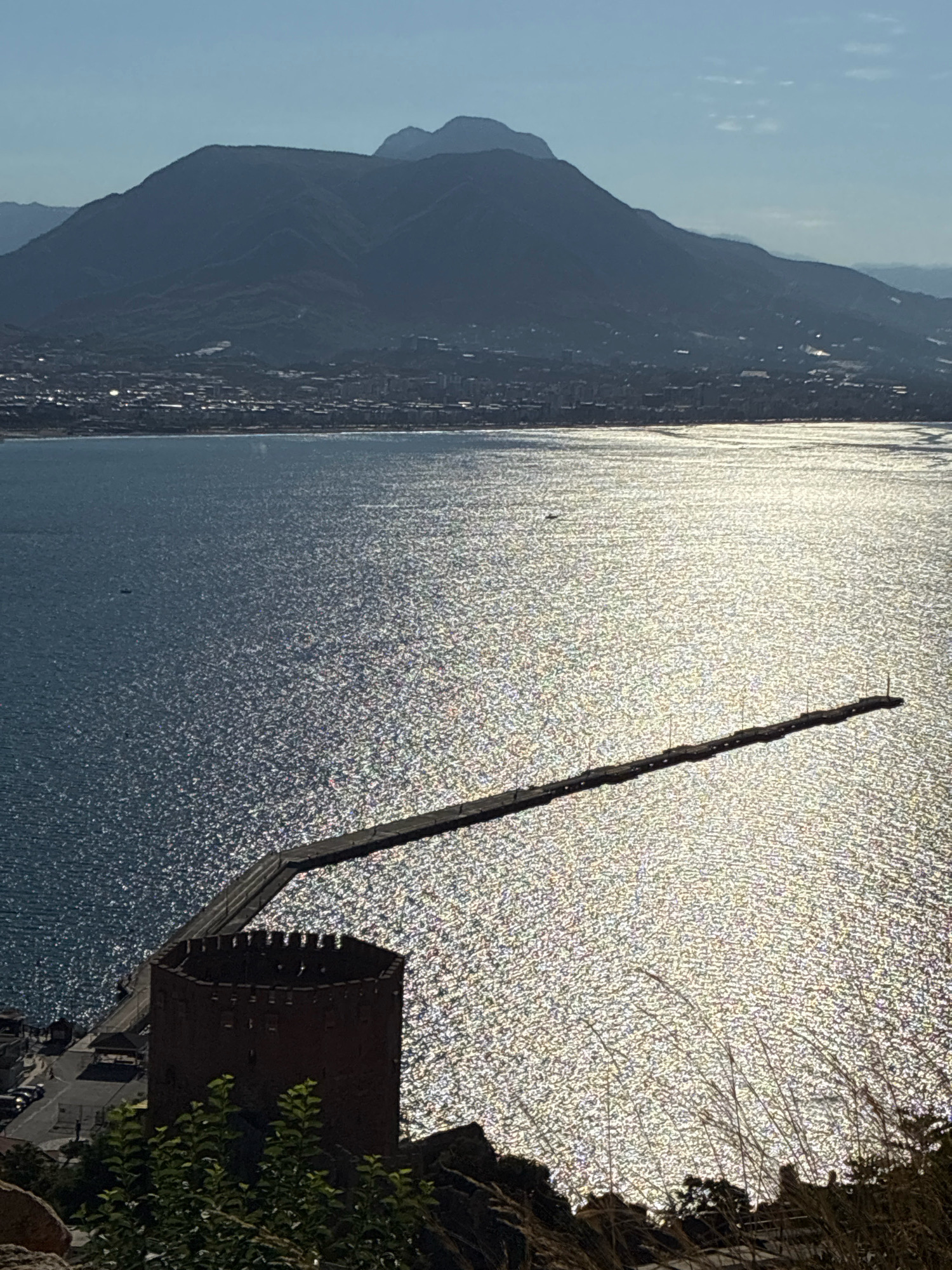 The image depicts a panoramic view from an elevated position overlooking the city of Alanya, Turkey. In the foreground, a section of a stone fortress, likely part of the Red Tower or a similar historical structure, is visible on the right. Behind the fortress, the Mediterranean Sea shimmers under the bright sunlight, with the surface of the water reflecting the light in a dazzling pattern.
Extending from the left side of the image and reaching into the sea is a long, dark pier or breakwater. This structure appears to be lined with poles, possibly for mooring boats or for illumination. The pier is situated in the middle ground of the image.
In the background, the city of Alanya sprawls along the coastline, nestled at the base of a range of imposing mountains. The mountains are silhouetted against a clear blue sky, their rugged forms softened by a light haze. The cityscape is a collection of buildings, some appearing to be residential, others possibly commercial or tourist-oriented, interspersed with roads and other urban infrastructure. The overall scene is bathed in the warm, bright light of a sunny day, with a slight haze contributing to the atmospheric perspective of the distant mountains.