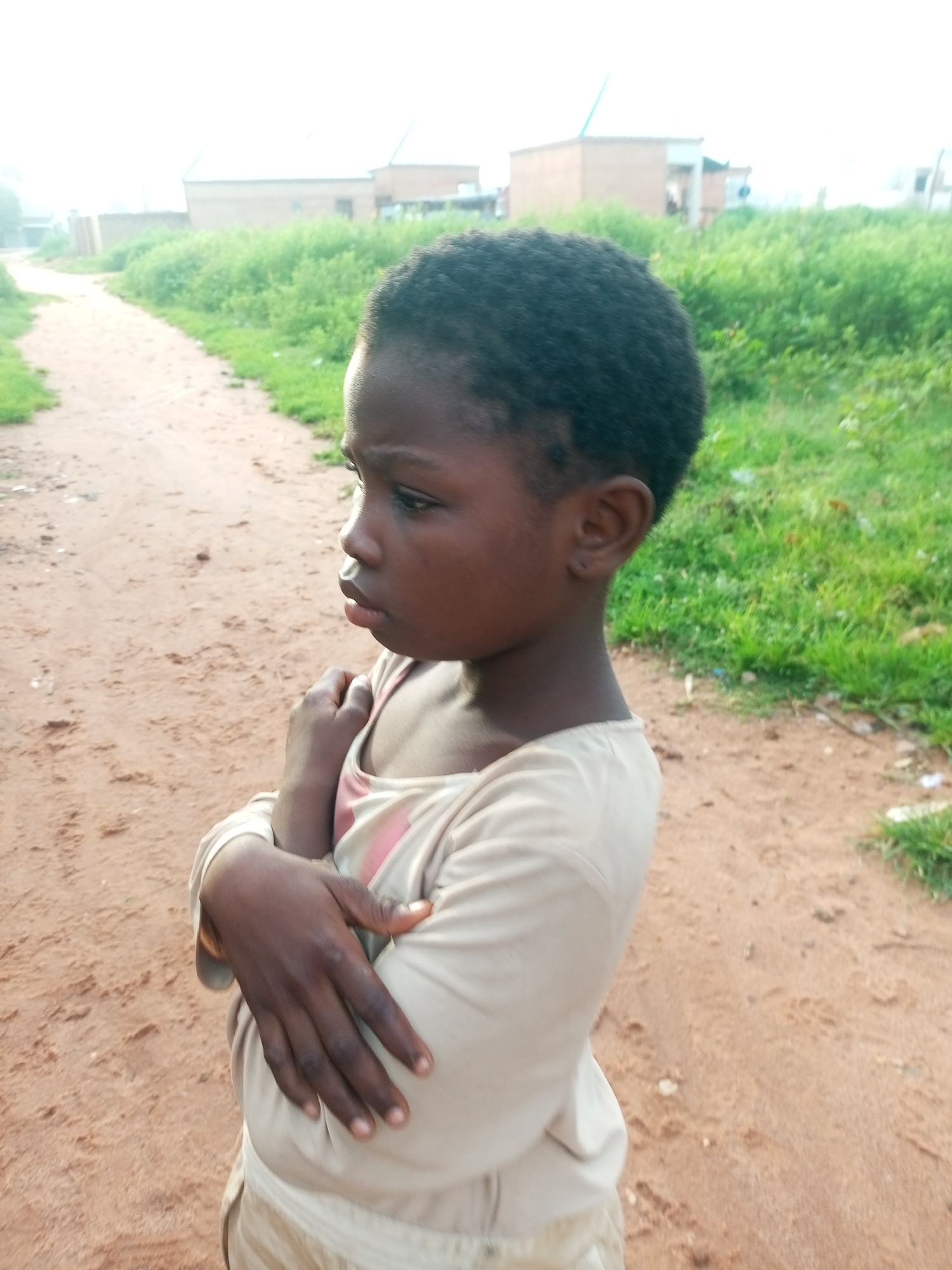 The image shows a young Black child with dark skin and short, dark hair, standing on a dirt path. The child is wearing a light-colored long-sleeved shirt with a pink stripe across the chest and light-colored pants. The child's arms are crossed over their chest, and they are looking to the left of the frame with a contemplative expression.
The background consists of a dirt path that curves to the left, lined with green vegetation on both sides. In the distance, there are several low-rise buildings with beige and white walls. The sky is bright and overcast. The overall impression is of a rural or semi-urban setting in Tempure, Nigeria. The lighting suggests it is daytime, possibly late afternoon or early morning due to the soft light.