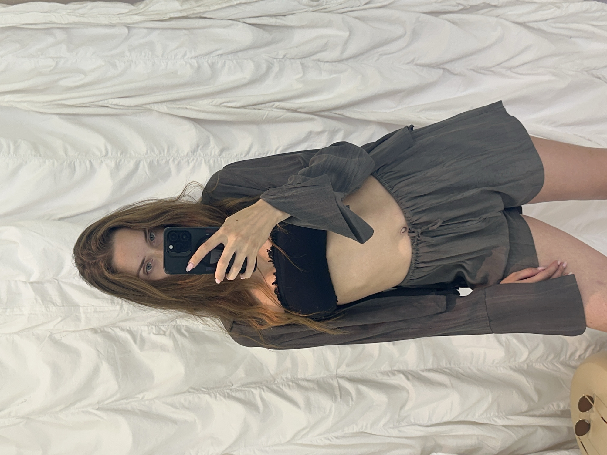 The scene, located in North Kuta, Indonesia, depicts a young adult female taking a mirror selfie indoors. She has long, light brown hair and light-colored eyes, partially obscured by a black smartphone held in her right hand. She is wearing a black strapless bandeau top with a ruffled upper edge, a dark grey or charcoal-colored long-sleeved, collared shirt worn open, and matching dark grey shorts with a drawstring waist. The clothing appears to be made of a textured or crinkled fabric. A belly button piercing is visible. Her fingernails are painted light pink. In the bottom left corner, a portion of a light-colored surface, possibly a stool or table, is visible with a pair of round, mirrored sunglasses resting on it. The background consists of a white or off-white fabric, possibly a curtain or wall hanging, displaying vertical folds and creases.