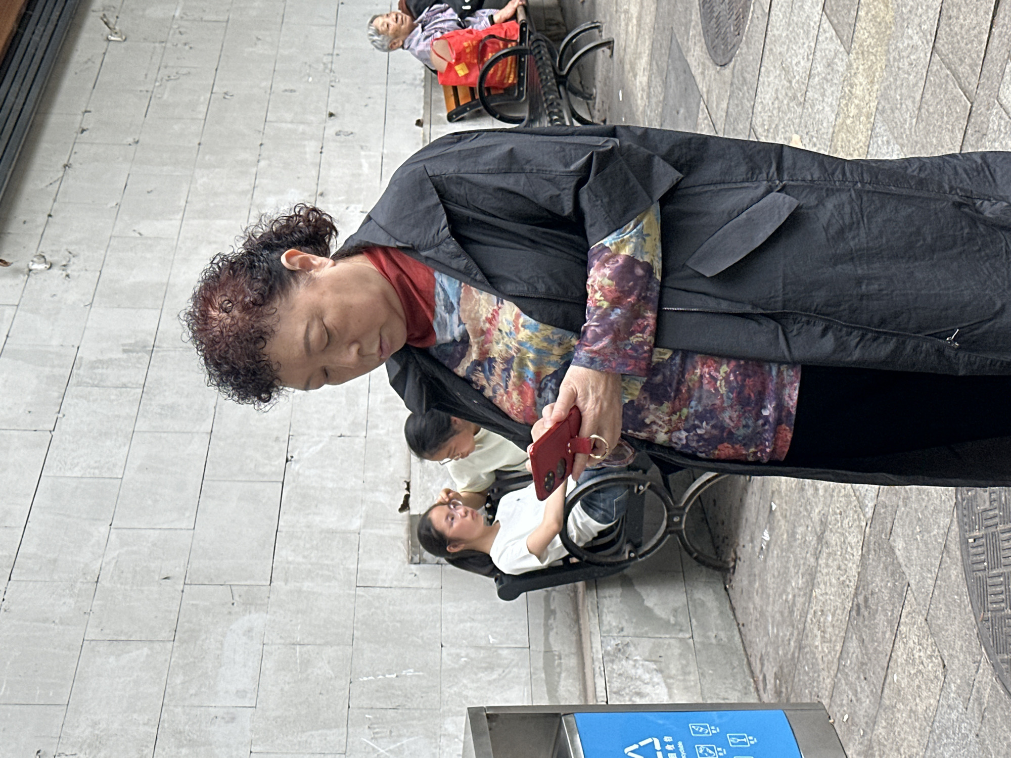 An image shows an elderly Asian woman standing outdoors and looking at a red smartphone. She is wearing a black jacket and a multi-colored floral shirt. Behind her, two younger Asian women are seated in wheelchairs. To the left is a gray wall. The ground is paved with rectangular stones. In the distance is an elderly person seated on a bench. There is a recycling bin at the bottom of the image. The location is in the city of Shapingba District, China.