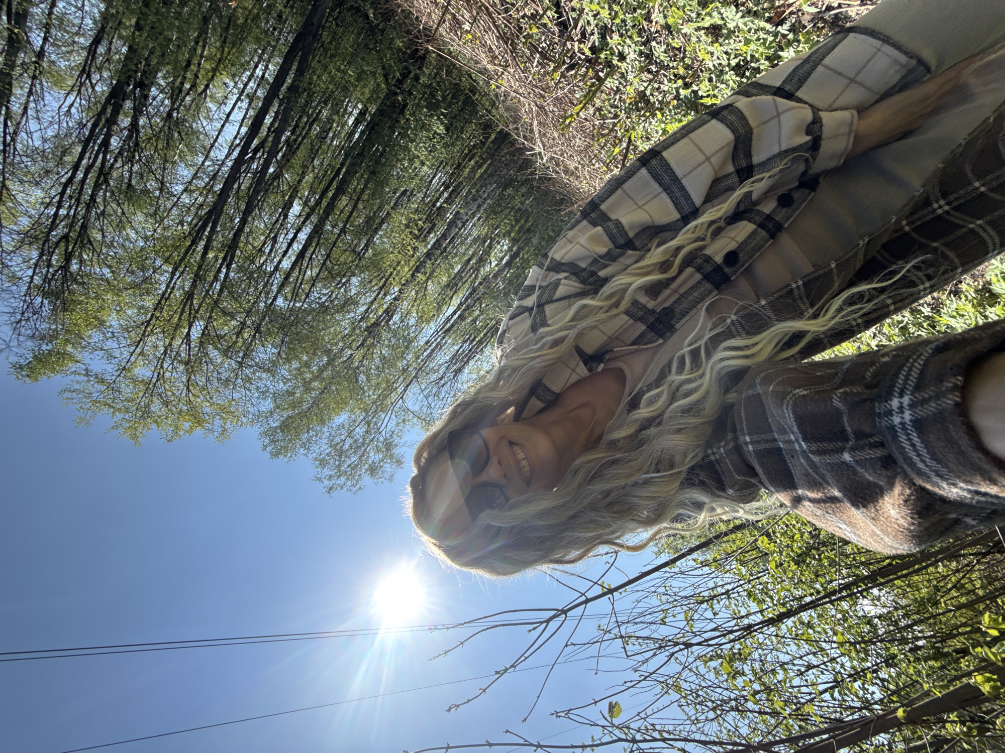 The image is taken from a low angle, looking upward at a woman wearing a plaid shirt and sunglasses. She is smiling at the camera. The background consists of trees, branches, and a bright blue sky with the sun shining through. The image is taken during daylight hours. Power lines are visible against the blue sky. It is not possible to determine that the location is in the city of Rusanovo, Russia.