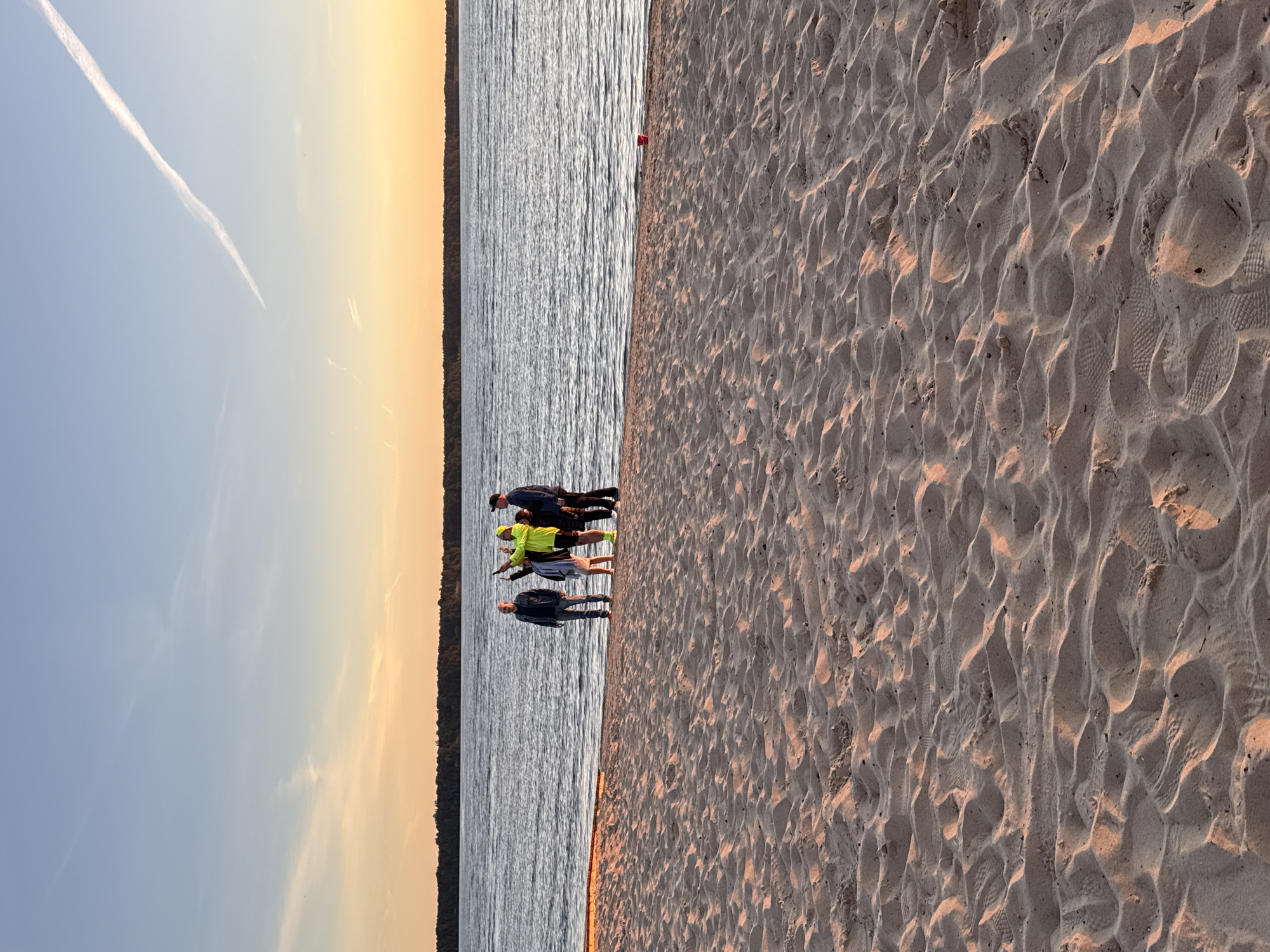 This image depicts a group of four individuals standing on a sandy beach at the edge of a body of water. The sky above is transitioning from a light blue at the top to a warm orange and yellow hue near the horizon, suggesting it is either sunrise or sunset. A faint contrail is visible in the upper portion of the sky. The water is calm with gentle ripples. A dark line of trees forms the distant shoreline. The foreground is dominated by the textured surface of the sand, showing footprints and undulating patterns. The group consists of two men in the front, one wearing a dark jacket and jeans, and another in a bright yellow jacket and lime green shoes. Behind them, a woman in a light-colored outfit and another man in a dark jacket are partially visible. One individual appears to be gesturing upwards. The overall scene is serene and bathed in the warm light of either dawn or dusk. There is no discernible indication in the image itself that the location is Tallinn, Estonia.