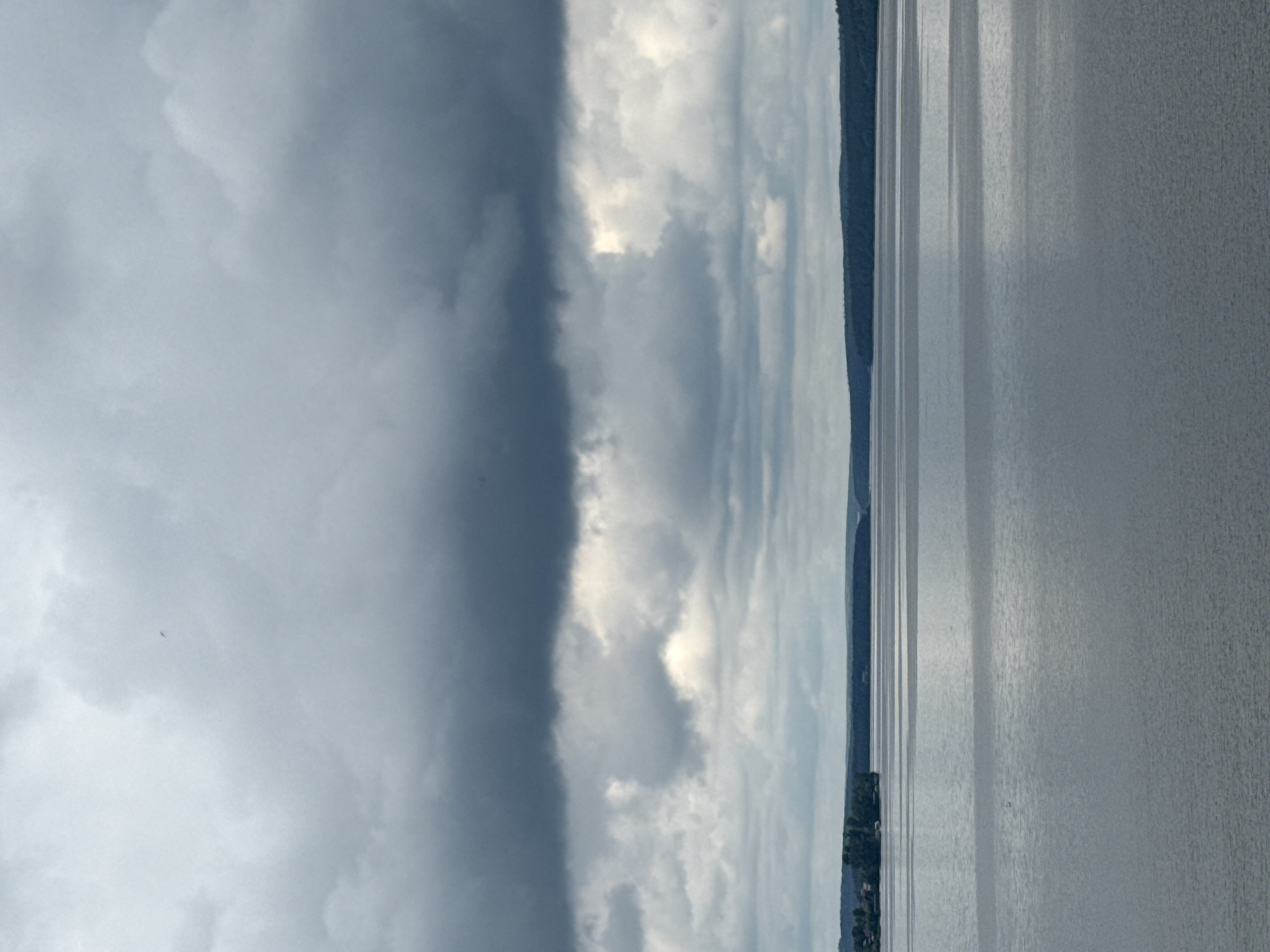 A wide landscape view features a large body of water extending across the foreground, meeting a low horizon line of distant land, and topped by a dramatic sky.
The water, likely a lake or reservoir, appears calm and reflects the overcast sky, displaying subtle variations in light and texture. A thin, dark strip of land, possibly hills or mountains, stretches across the mid-ground horizon, becoming slightly more pronounced on the right side where a small cluster of trees or buildings can be discerned.
The sky occupies the upper portion of the image, showcasing a striking contrast in cloud formations. The left side is dominated by heavy, dark gray clouds, suggesting a storm front or dense cloud cover. Towards the right, the clouds lighten, with brighter, diffuse sunlight breaking through, illuminating patches of the sky and creating a more dynamic appearance.
The scene captures a moment of changing weather, possibly an approaching or receding storm, characterized by the dramatic interplay of light and shadow in the clouds. The time of day is ambiguous but suggests either early morning or late afternoon, given the diffused light. No specific activity or individuals are visible. There is no visible text. Given the context, the location is likely the Votkinsk Reservoir in Votkinsk, Russia.