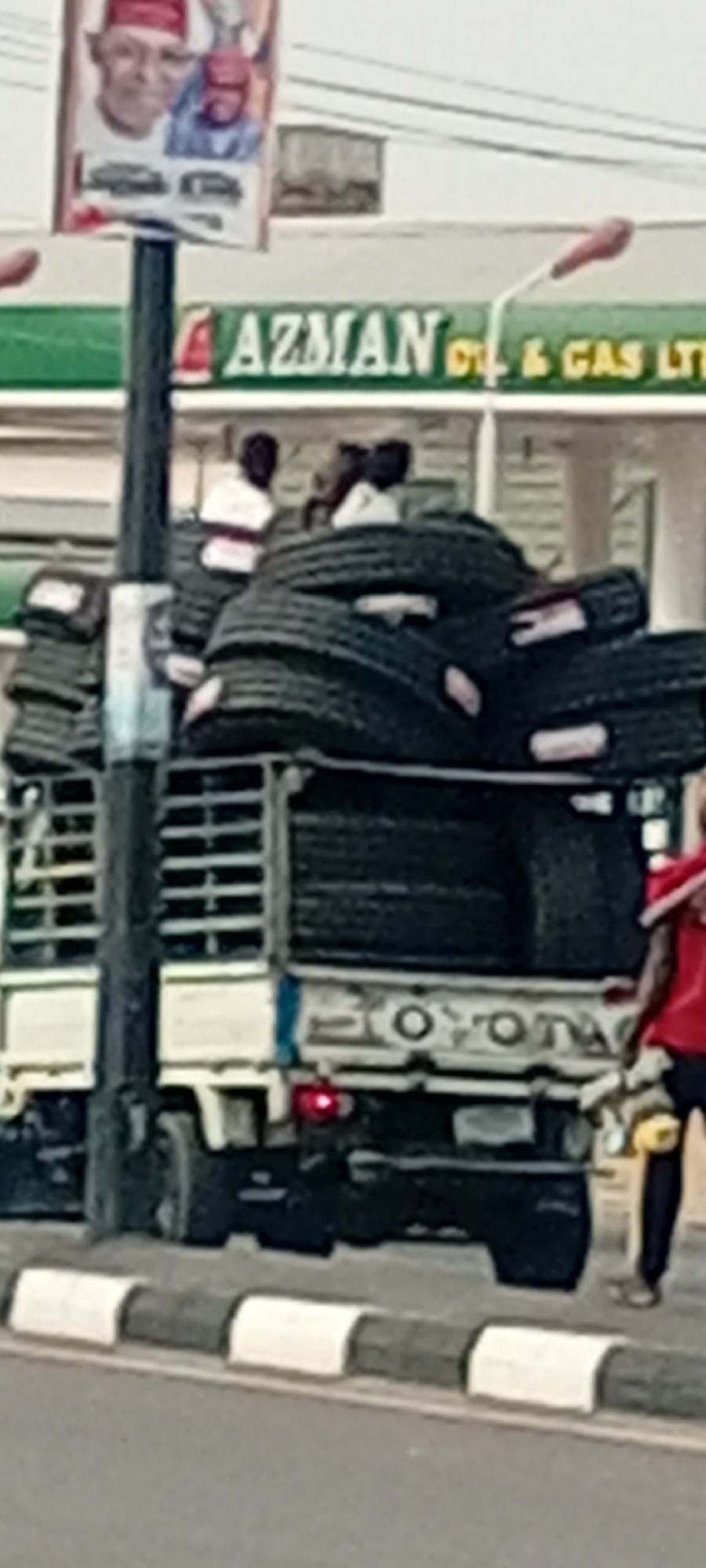 A white Toyota truck is overloaded with tires, obscuring the truck bed. Two people are riding atop the tires. A man wearing a red shirt stands beside the truck, holding yellow cans. The scene is located in Tudun Wada, Nigeria, as indicated by the "AZMAN Oil & Gas LTD" sign and a political poster featuring individuals wearing red hats. The truck is parked alongside a street curb painted with alternating black and white segments.