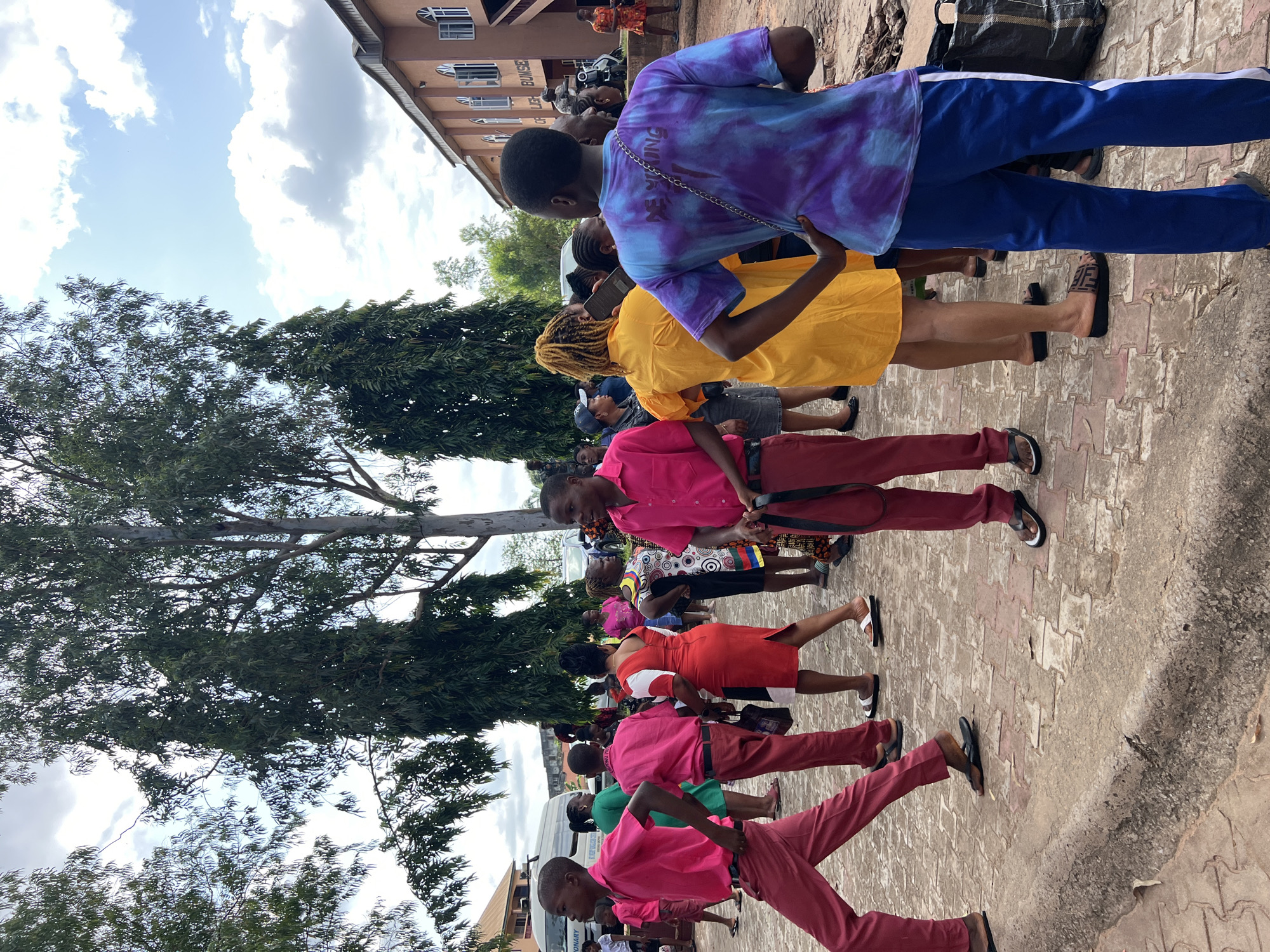 In Nnewi, Nigeria, a group of people are gathered outside a building with a light brown facade and arched windows. The building appears to be a place of worship or a similar institution, as indicated by text visible on its exterior. A tall, leafy tree dominates the left side of the frame, its branches extending towards a cloudy sky. The ground is paved with patterned bricks.
In the foreground, several individuals are seen. A young man on the far left, facing left, wears a bright pink long-sleeved shirt and matching red pants. He walks with a dynamic stride, his left leg extended. To his right, another young man in a similar pink shirt and red pants faces away from the camera, looking towards the right. He holds a dark bag in his left hand. Further into the crowd, a woman in a red dress with white and black panels walks away from the camera, her back to the viewer.
A person in a bright yellow dress, with braided blonde hair, stands facing away from the camera, holding a dark object, possibly a phone or book. To their right, a young person in a tie-dye t-shirt with purple and blue hues, and blue pants with a white stripe down the side, stands with their hands on their hips, looking to the right. Their t-shirt has writing on the back.
Other individuals are visible in the background, some in brightly colored attire, suggesting a gathering or event. Vehicles, including a white van, are parked in the vicinity. The overall scene depicts a lively outdoor gathering in a semi-urban environment.