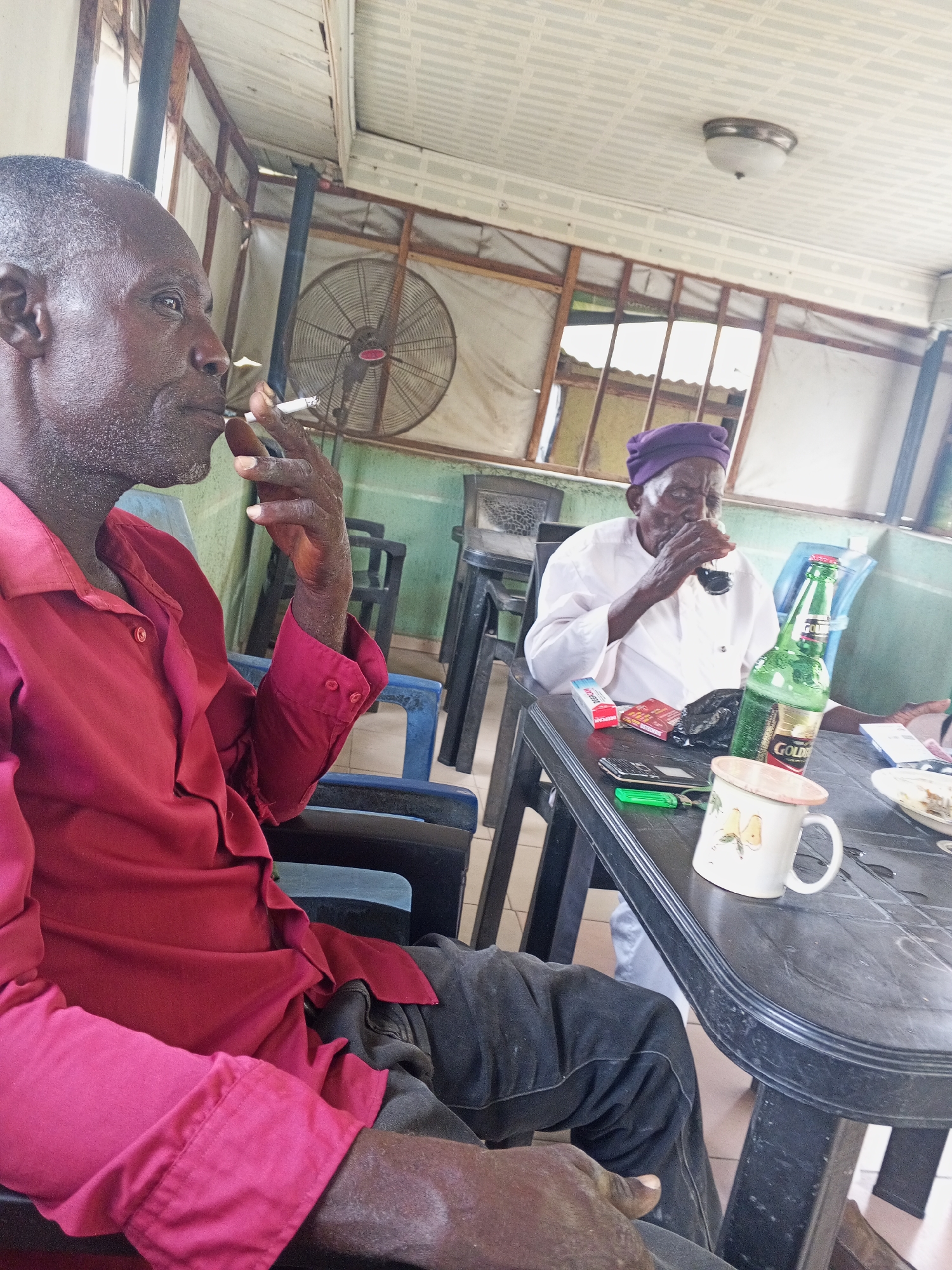 The image shows two older African men in a modest, partially open-air establishment. The location is assumed to be Zango-ndaji, Nigeria.
One man, wearing a red button-down shirt, sits to the left of the frame and is smoking a cigarette. The other man, wearing a white top and a purple cap, is seated at a black plastic table. He is drinking from a glass. On the table, there is a bottle of "GOLDEN" beer, a mug, a pack of cigarettes, a cell phone, a green lighter and what appears to be a plate with food. A large fan is visible in the background. The building has a partial ceiling and open window spaces with a wooden frame.