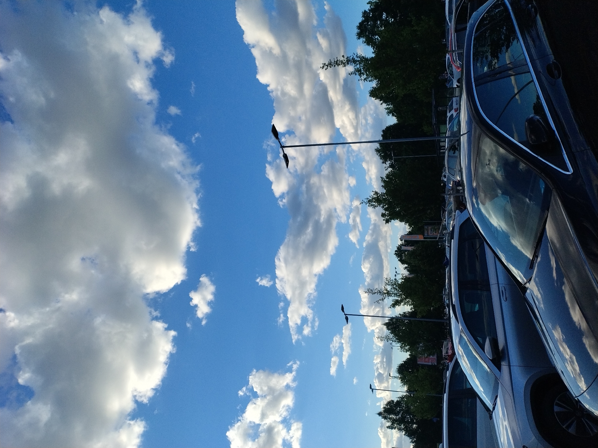The image shows a parking lot in Písek, Czechia, under a bright blue sky filled with fluffy white clouds. Several cars are parked, including a silver SUV in the foreground. Tall streetlights with dark lamps are visible, along with a line of dark green trees behind the cars. In the background, some buildings and flagpoles can be seen. The scene appears to be during daylight hours, with the sun casting reflections on the car surfaces.