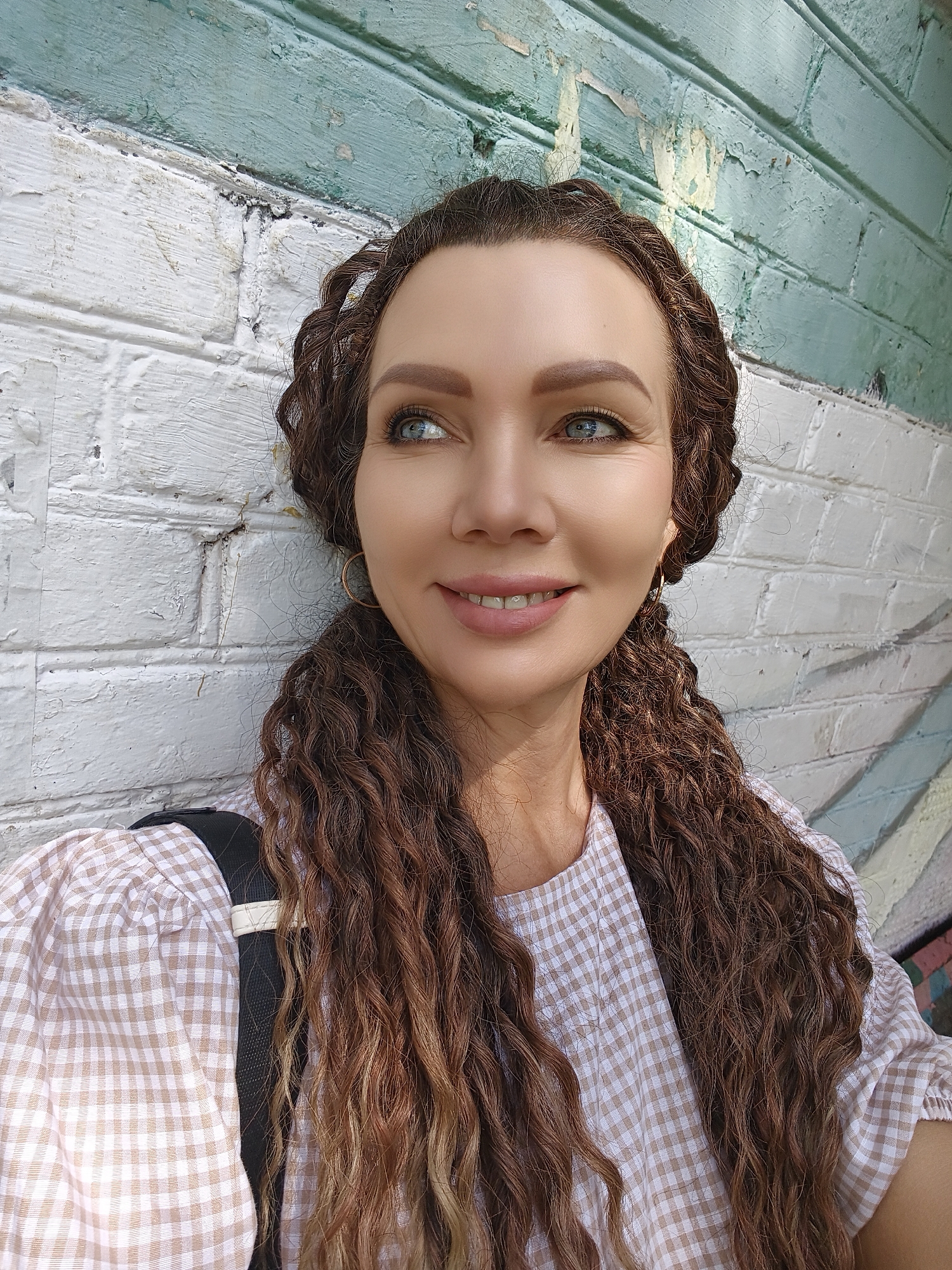 A woman is photographed from the chest up, leaning against a brick wall. The wall is painted white and has a section of peeling teal paint at the top. The woman has long, dark curly hair, with some strands pulled back into braids. She is wearing a light-colored, checkered shirt. Her eyes are blue and she is looking to the side with a slight smile. A backpack strap is visible over her shoulder. The background wall shows signs of graffiti art with colorful markings.