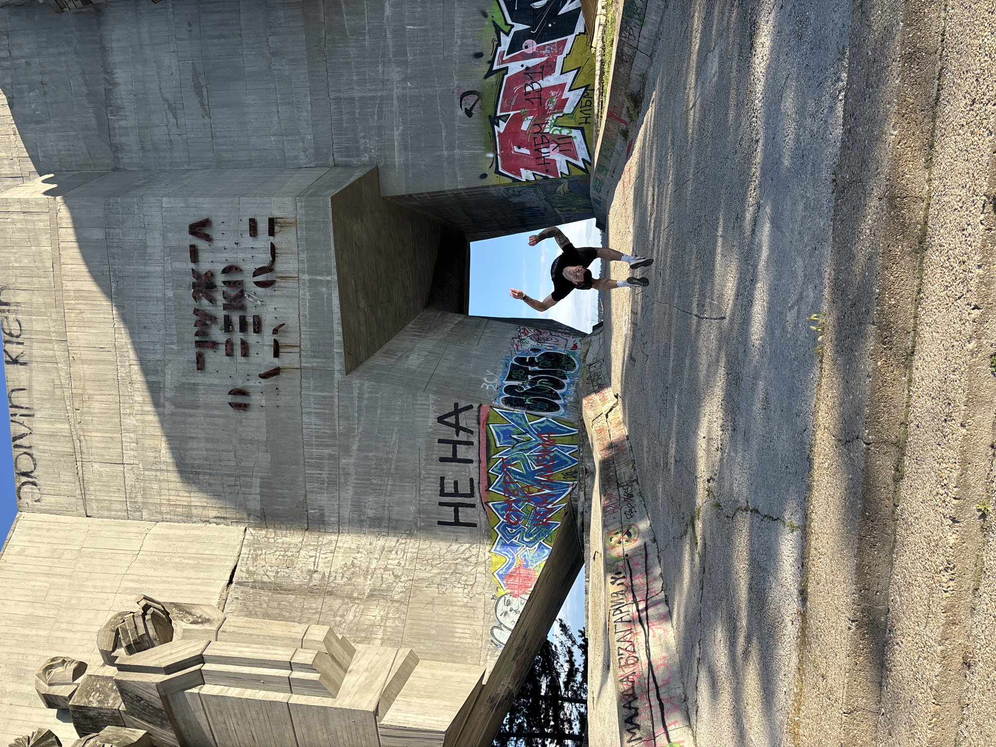 The image shows a person standing in the middle of a concrete structure covered in graffiti. The structure appears to be a bridge or tunnel, with a rectangular opening in the distance. The person is wearing dark clothing and white socks. The ground is concrete with weeds growing along the edge. The graffiti includes various tags and drawings in different colors. The location is Varna, Bulgaria, based on the graffiti on the structure.