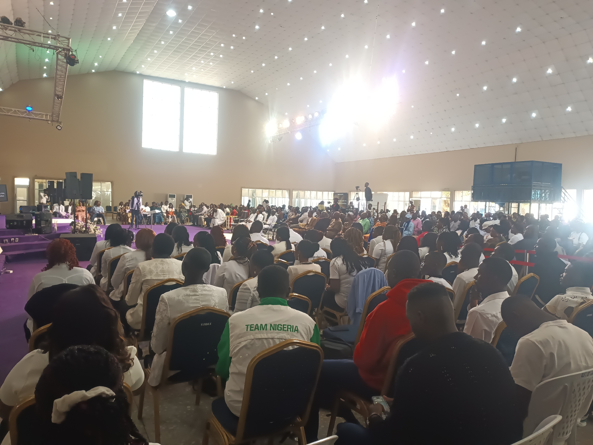 The image captures an indoor gathering of a large audience within a spacious hall. The hall features a high, illuminated ceiling with numerous recessed lights. Two large, vertically oriented windows are prominent on the far wall, letting in a bright, natural light that creates a glare. On the left side of the image, a stage area is visible with truss lighting structures and large speakers. A camera on a tripod is positioned in the center of the hall, facing the audience. The audience members are seated on rows of chairs, facing a central point of focus, likely a stage or presentation area, which is not fully visible. Many individuals are wearing white or light-colored clothing. One person in the foreground is wearing a green and white vest with "TEAM NIGERIA" printed on the back. The overall scene suggests a conference, religious service, or a similar large-scale event. The location is identified as Gura Top, Nigeria.