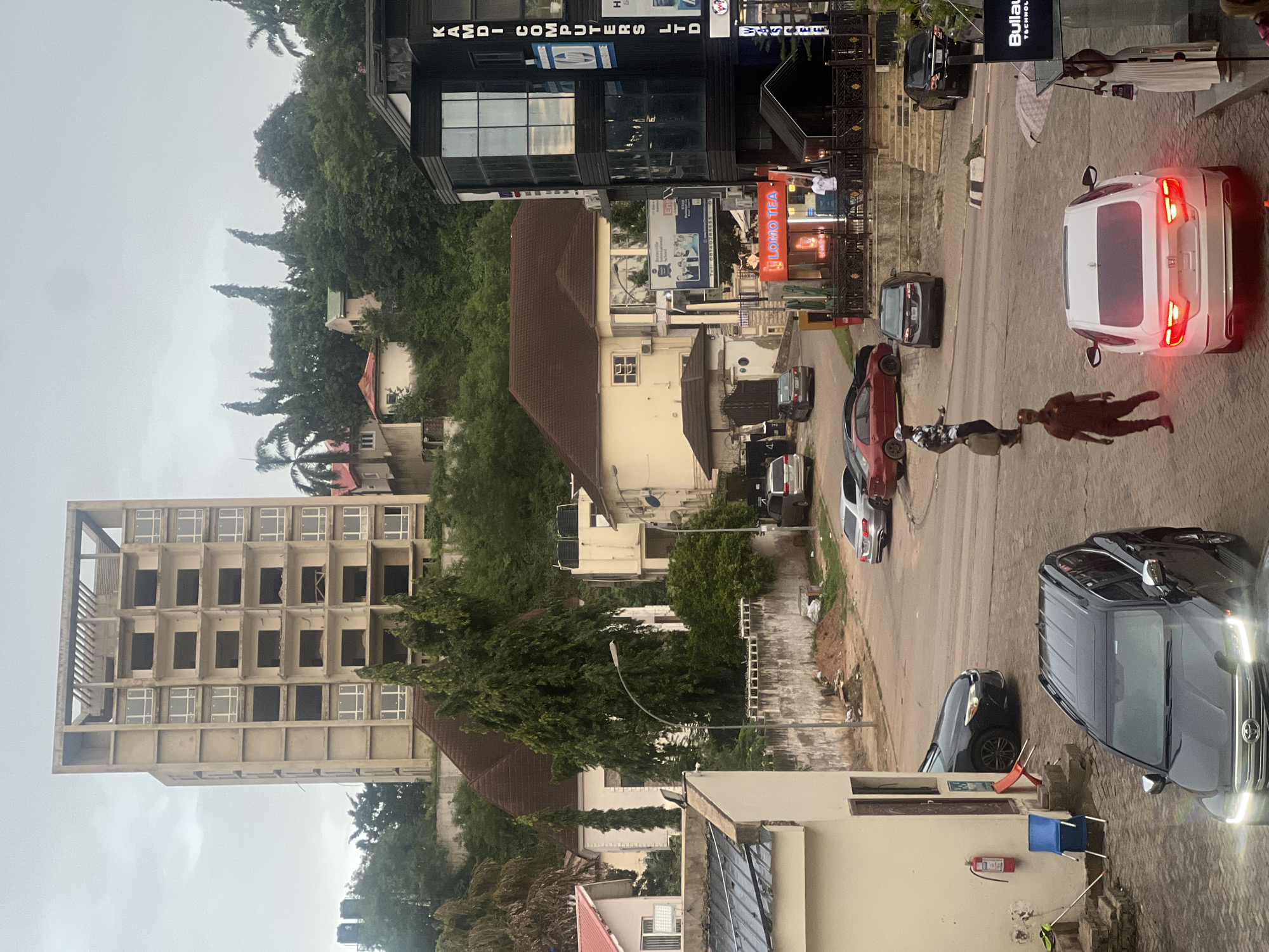 This is a street scene in Abuja, Nigeria, under an overcast sky. In the foreground, a dark gray SUV is parked, facing forward with its headlights on. To its right, a white sedan is stopped at an intersection, its brake lights illuminated. A person in a dark uniform walks away from the sedan. Further up the street, a man carrying a bag walks towards the left side of the frame.
Several cars are parked and moving on the street. A black sedan is parked near a building on the left. In the middle ground, a red sedan is parked. There is a multi-story building with a dark facade and glass windows on the right, displaying signage for "KAMDI COMPUTERS LTD." Below it, a smaller establishment with an orange sign reading "LOMO TEA" is visible.
In the background, a tall, unfinished concrete building with visible window frames stands on a hillside. Numerous trees and other buildings, some with red roofs, are situated on the sloped terrain. A fire extinguisher is mounted on the side of a light-colored building in the lower left corner, next to two blue chairs. The ground is paved with interlocking bricks.