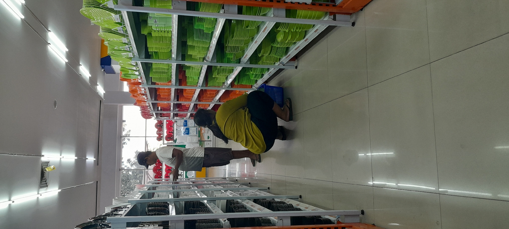 The image depicts the interior of a retail store selling household items, specifically tableware. In the foreground, a person wearing a yellow t-shirt and dark pants is squatting and looking at items on a shelf. To their left, a man in a white t-shirt and dark shorts stands and examines items on a shelf. The shelves are stocked with plates and bowls in various colors, predominantly green and orange. The store appears to be well-lit with fluorescent lights on the ceiling. The flooring is tiled. The scene is located in Rogojampi, Indonesia, within a retail establishment.