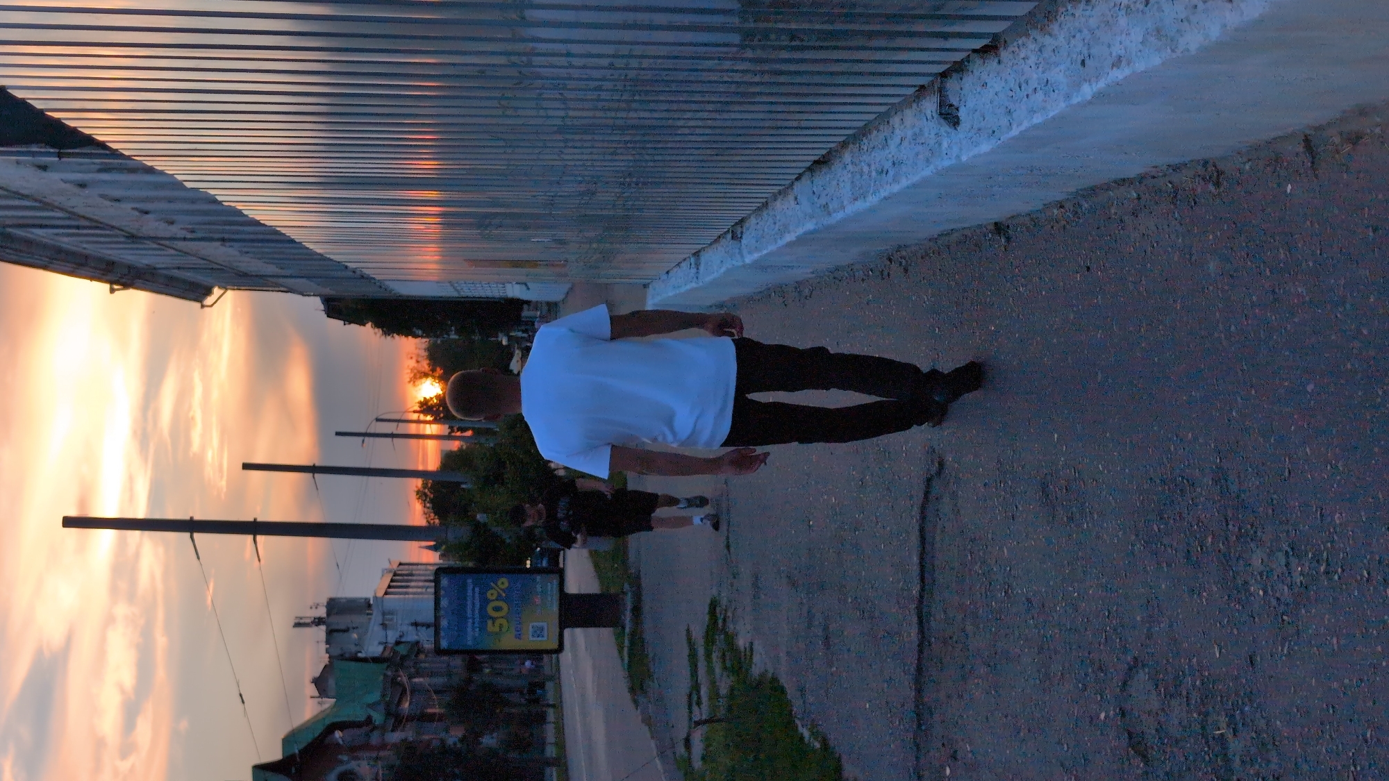 The image shows two individuals walking on a sidewalk during sunset in Starovirivka, Ukraine. The sky is filled with a dramatic orange and yellow sunset, with clouds catching the warm light. A corrugated metal fence runs along the left side of the frame, casting long shadows. The ground is paved with asphalt.
The foreground subject is a person wearing a white t-shirt and dark pants, walking away from the camera. Their posture is relaxed, suggesting a leisurely stroll. Behind them, another person in dark clothing is visible, also walking along the sidewalk.
In the background, trees and buildings are silhouetted against the vibrant sky. Streetlights are also visible, indicating a transition from daylight to evening. A digital advertisement panel is present on the sidewalk, displaying "50%" prominently, suggesting a sale or promotion. The overall mood is tranquil and serene, characteristic of a sunset scene. The time of day is clearly dusk.