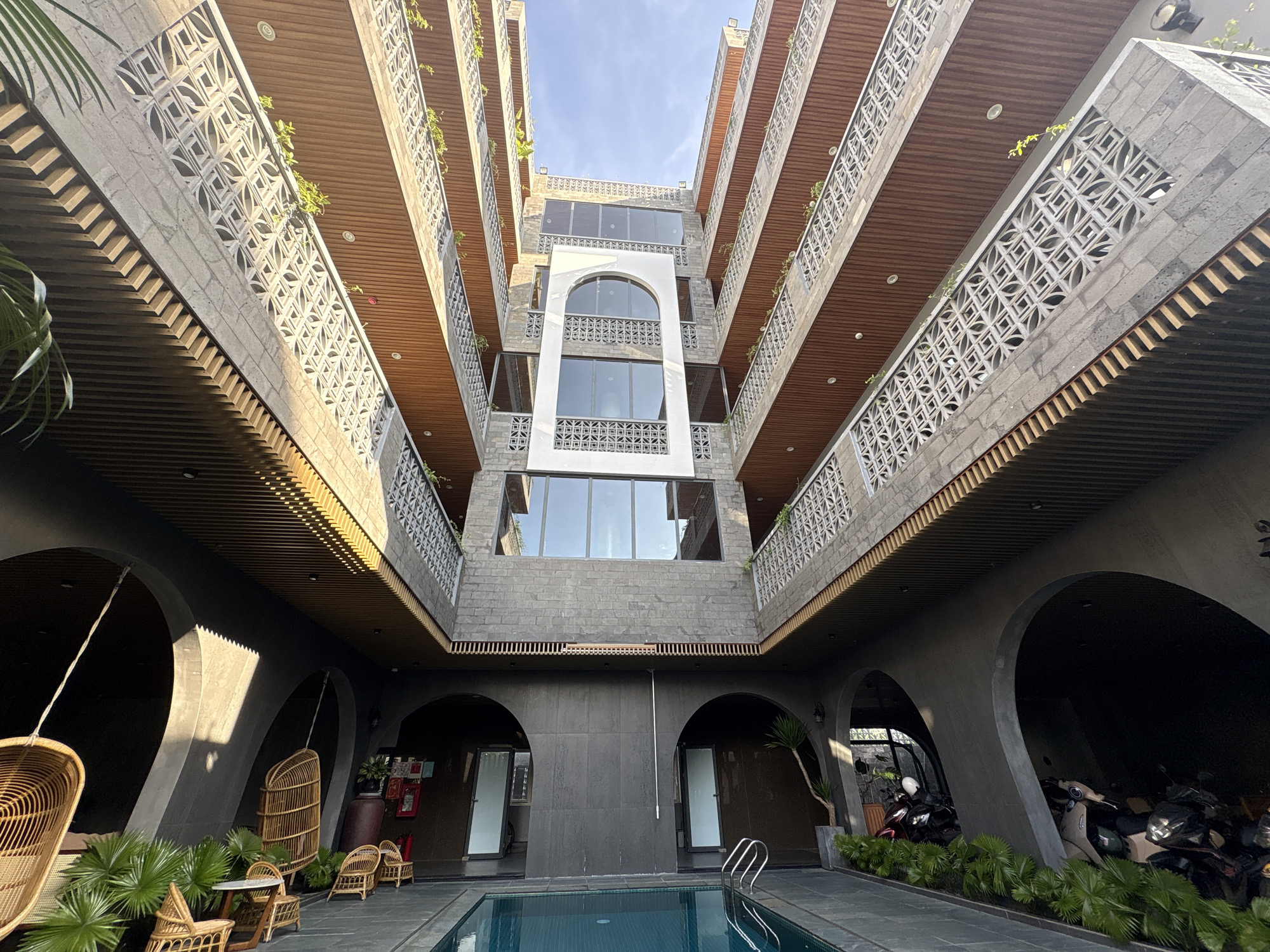 The photograph showcases the interior courtyard of a building, potentially a hotel or residential complex, located in the city of Đà Nẵng, Vietnam. The structure features a distinctive architectural design with multiple levels of balconies and walkways that overlook a central swimming pool. The building's exterior is composed of gray stone or concrete, accented by wooden undersides of the balconies and white, decorative balustrades. Arched openings are present at the ground level, leading into the building's interior.
The swimming pool is the focal point of the courtyard, surrounded by gray stone paving. Near the pool, there are lounge areas with wicker chairs and hanging cocoon-like seats, suggesting a relaxation and leisure space. Tropical plants add greenery to the environment, softening the hard lines of the architecture.
In the background, motorcycles are visible parked under one of the arched entryways, providing a sense of everyday life in the space. The sky is visible through the top of the courtyard, indicating an open-air design. The overall atmosphere is one of modern luxury combined with tropical elements.