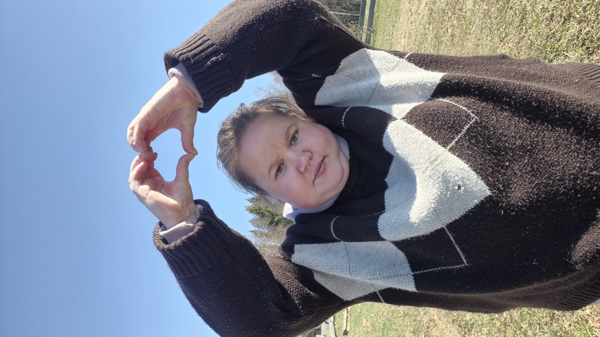 A woman with fair skin and blonde hair is outdoors under a clear blue sky, forming a small heart shape with her fingers. She is wearing a brown and white argyle sweater. The background consists of a grassy area with some trees visible in the distance. The location is Дойная, Russia.
