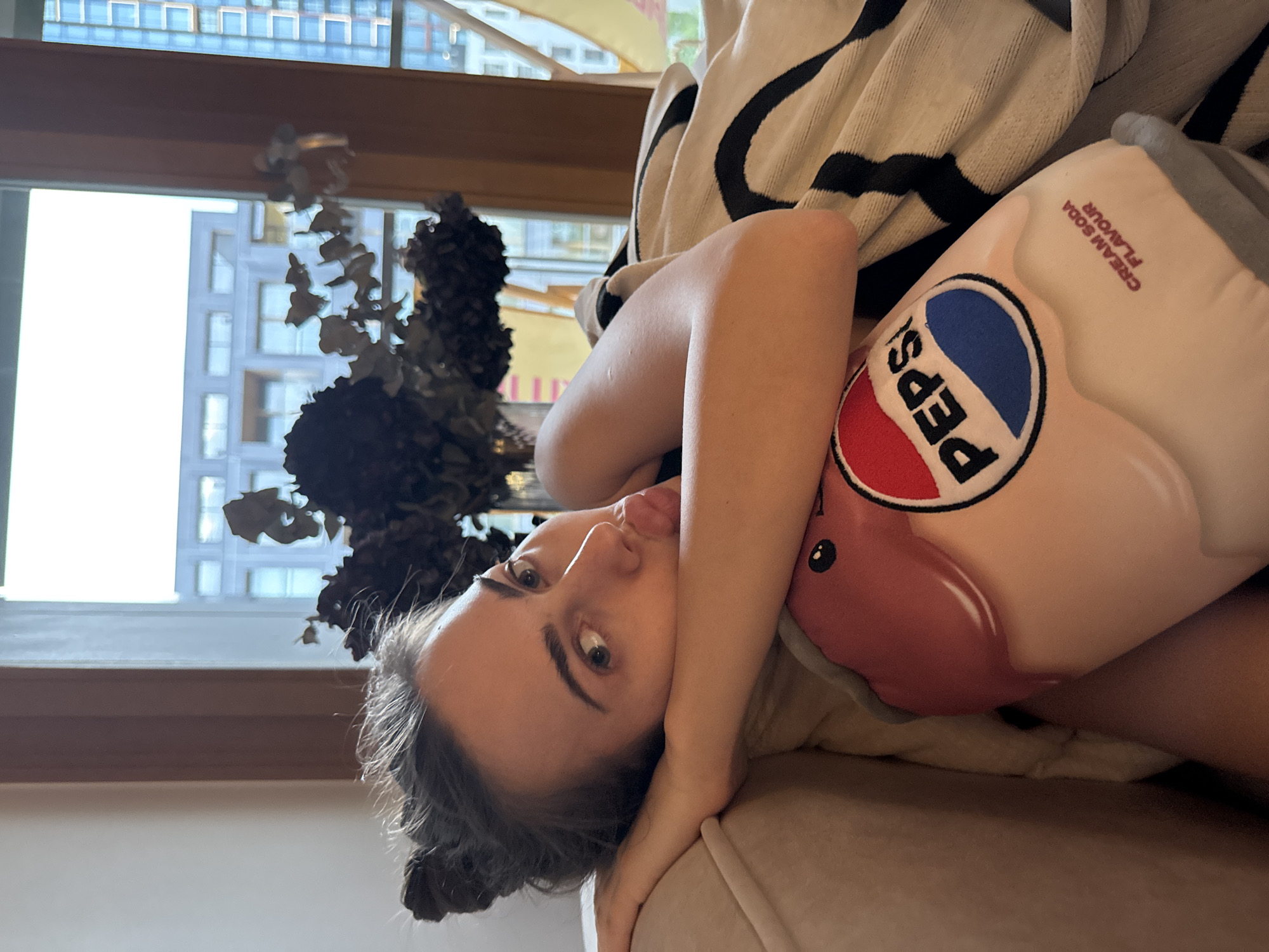 A young woman is depicted lying on her side, looking directly at the camera, in what appears to be an indoor setting like a living room or bedroom. She has dark hair tied back and her facial expression is neutral to slightly playful, with her tongue peeking out. She is resting on a light-colored surface, possibly a couch or bed, and is partially covered by a striped blanket or throw with black and white patterns.
The woman is hugging a large, soft, light-pink plush toy shaped like a Pepsi can. The plush prominently features the classic red, white, and blue Pepsi logo, and text clearly visible reads "CREAM SODA FLAVOUR".
In the background, a large window with a wooden frame allows bright daylight to enter, illuminating the scene. Through the window, modern, tall buildings are visible, indicating an urban environment. To the left side of the frame, near the window, there is a dark, possibly dried, floral arrangement or plant. The overall impression is a relaxed, daytime moment indoors.