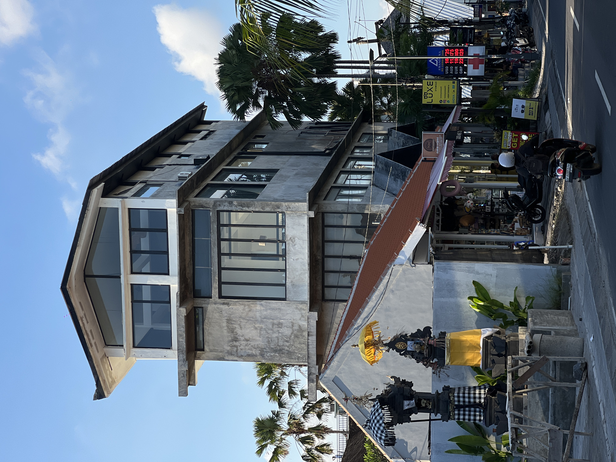 The image depicts a street scene in North Kuta, Indonesia. A modern four-story building with a concrete facade and large windows dominates the left side of the frame. A palm tree is visible behind the building.
On the right side of the image, a row of shops and restaurants lines the street. Several signs advertise businesses, including an eyewear store called "LUXE" and an exchange rate board listing prices for USD, AUD, EUR, and GBP. A person is riding a motorcycle on the street. Traditional Balinese shrines with black and white checkered cloths and yellow offerings are positioned near the storefronts. The sky is blue with scattered clouds.
