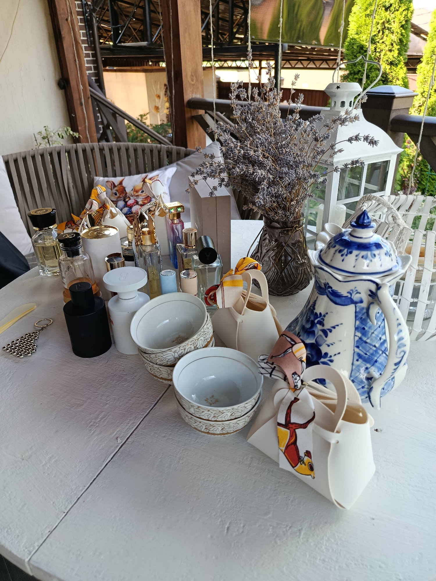 A close-up shot displays a collection of assorted items arranged on a white, distressed wooden table, possibly located outdoors. In the foreground, several white ceramic bowls with gold trim are stacked, with one prominently visible. Adjacent to them, a decorative white teapot with blue floral patterns sits on the table. To the left, a cluster of perfume bottles of various shapes and sizes, some with gold caps, are scattered across the surface. A black cylindrical bottle and a white bottle with a rounded cap are also present. A keychain with a decorative silver charm rests near the edge of the table. Behind these items, a bouquet of dried lavender in a dark vase is positioned next to a white lantern with a star-shaped cutout. In the background, a rustic wooden porch structure with exposed beams and a thatched roof can be seen. The environment suggests a relaxed, possibly domestic, setting. The location in Misaylovo, Russia, is noted, though not visually evident in the provided image. There are no people visible in the scene.