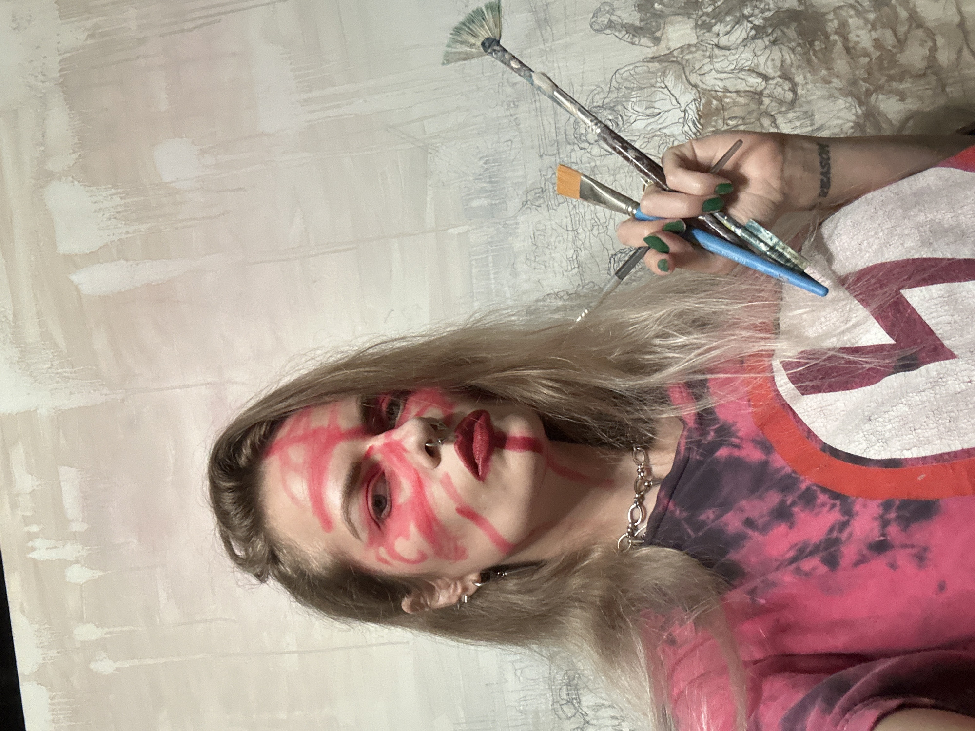 A woman is pictured indoors, likely at an art studio, holding three paintbrushes in her right hand. She has blonde hair and fair skin with red paint smeared across her face. She is wearing a tie-dye shirt, a silver chain necklace, and earrings. Her fingernails are painted green, and a tattoo is visible on her wrist. In the background, there is a partially painted canvas with brushstrokes and a faint drawing. The location cannot be determined to be in Cape Coral, Florida, or any specific location.