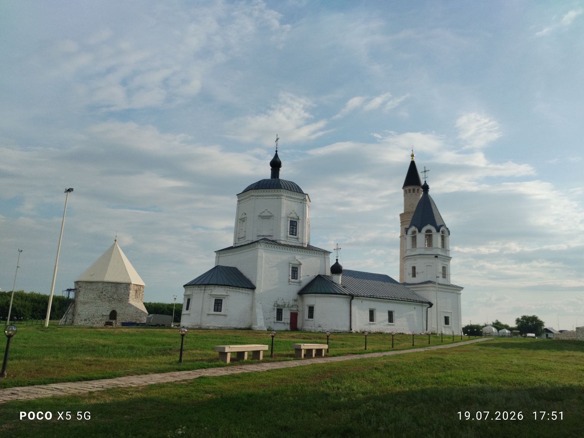 The image shows the White Chamber (also known as the Church of the Assumption of the Blessed Virgin Mary) and the Khan's Mosque in the city of Bolgar, Russia. The White Chamber is a prominent white brick building with a dark pitched roof and a single dark dome topped with a cross. To its right is the Khan's Mosque, a taller, multi-tiered white structure with a dark, conical roof and a cross at its apex. To the left of the White Chamber is a conical stone building with a lighter roof, likely the remnants of an older structure. The buildings are situated on a grassy landscape with a paved walkway leading towards them. Several lampposts are visible along the walkway, and two light-colored benches are placed beside it. In the background, trees and some white tent-like structures can be seen. The sky is partly cloudy. The bottom of the image displays a watermark reading "POCO X5 5G" on the left and "19.07.2026 17:51" on the right, indicating the date and time the photograph was taken. There are no people visible in the scene.