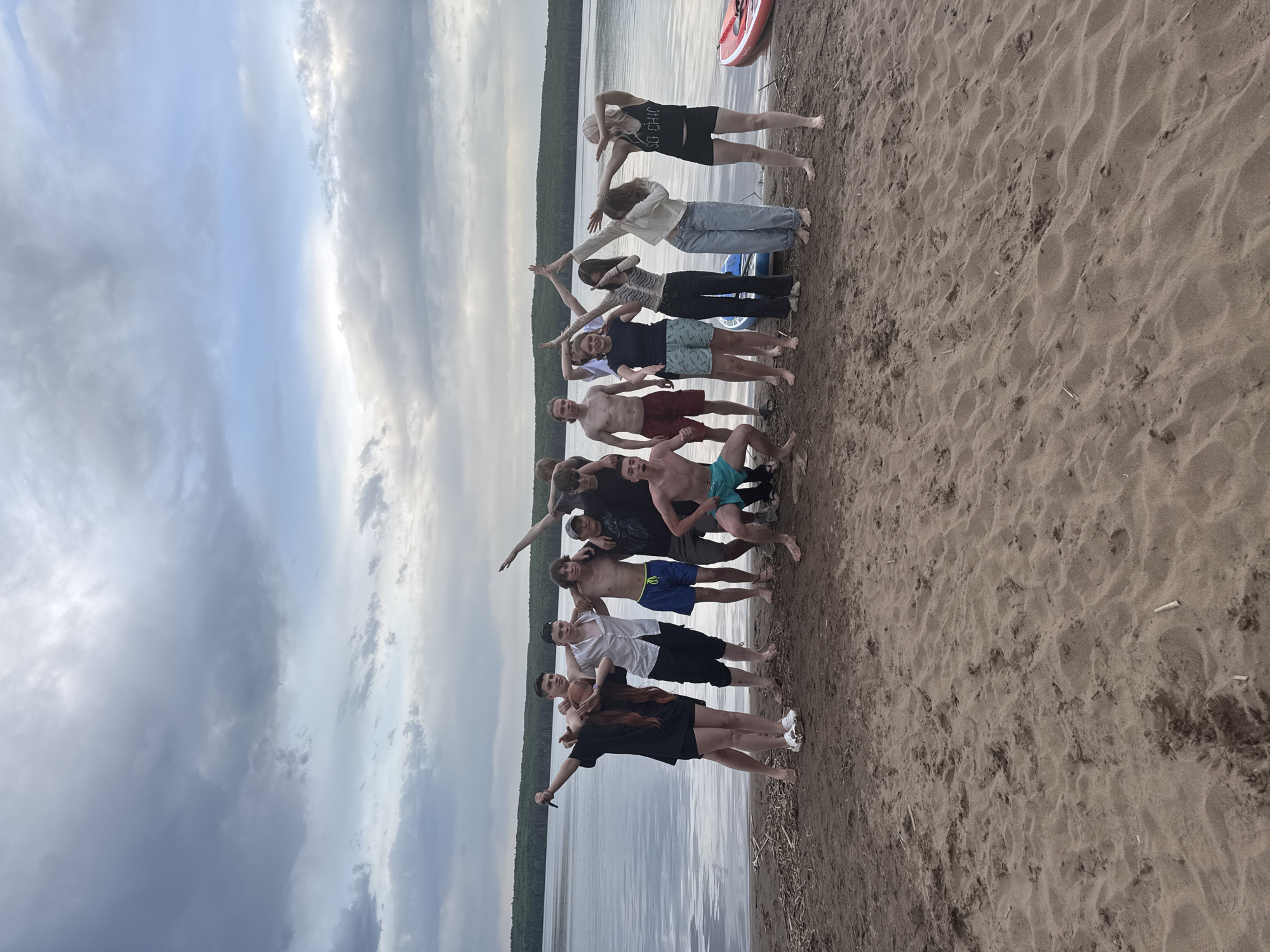A group of about 13 people is standing on a sandy beach along a body of water, likely a lake or river. They are posed to form the letter "T" with their bodies, with some people holding their arms out to complete the shape. The sky is cloudy. The location is likely Chaikovsky, Russia, as specified in the prompt. The water appears calm, and there is a treeline visible on the opposite shore. There are a few objects, possibly kayaks, on the beach near the group. The group appears to be a mix of genders and ages. Some are wearing swimsuits, while others are in casual clothing. They appear to be interacting in a playful or celebratory way. The beach is sandy and appears to be public.