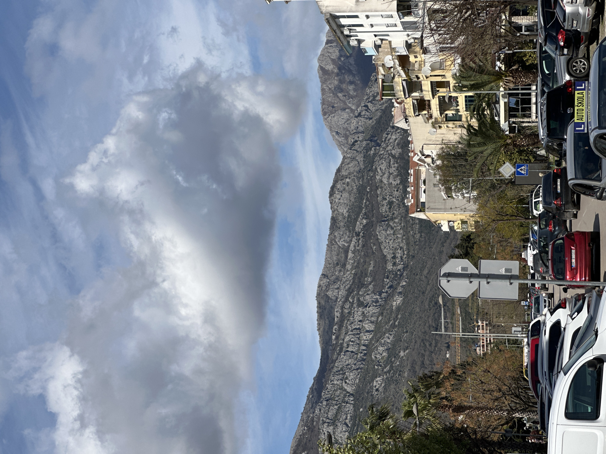 The image captures a scene in Šušanj, Montenegro, featuring a juxtaposition of the natural landscape and urban elements. A prominent mountain range dominates the backdrop, contrasting with the buildings and structures along a city street. Vehicles are parked along the side of the road, and street signs are visible. The sky is partly cloudy.