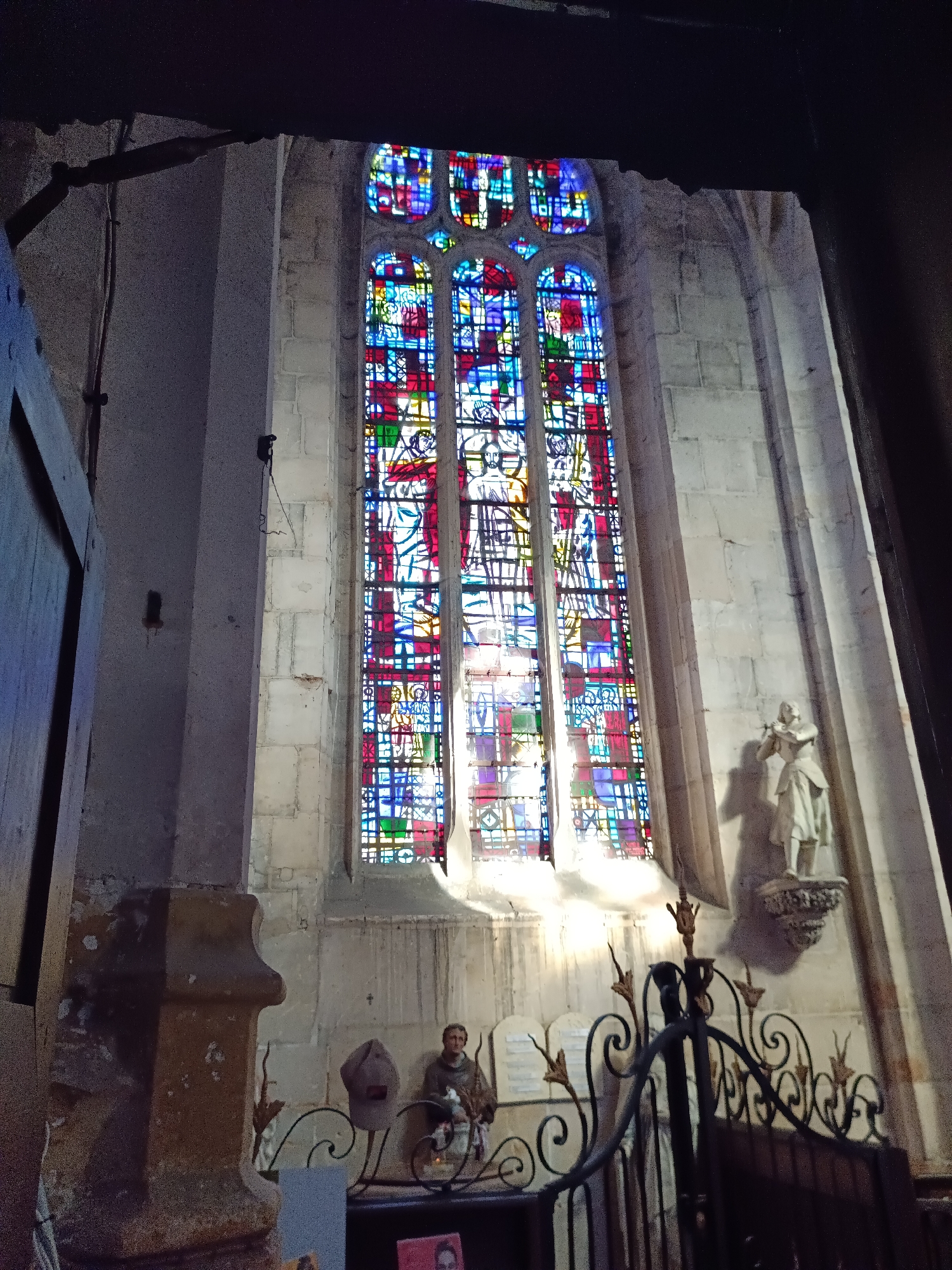 The image shows the interior of the church of Saint-Pierre in Le Neubourg, France. The primary focus is a tall, narrow stained-glass window with a Gothic arch, filled with a colorful geometric and figurative pattern. The window is illuminated from behind, casting colored light onto the stone wall. To the right of the window, on a small pedestal, stands a white statue of a person in historical attire, possibly a saint or historical figure. In the foreground, a dark wrought-iron gate with decorative scrollwork separates the viewer from a lower section of the church. Below and to the left of the stained-glass window, near the gate, are two small statues, one appearing to be a monk seated and the other a bust with a hat. To the far left, a portion of a large wooden door or architectural element is visible. The surrounding architecture consists of rough-hewn stone blocks, characteristic of medieval construction. The lighting is dim, with the stained-glass window being the brightest element.