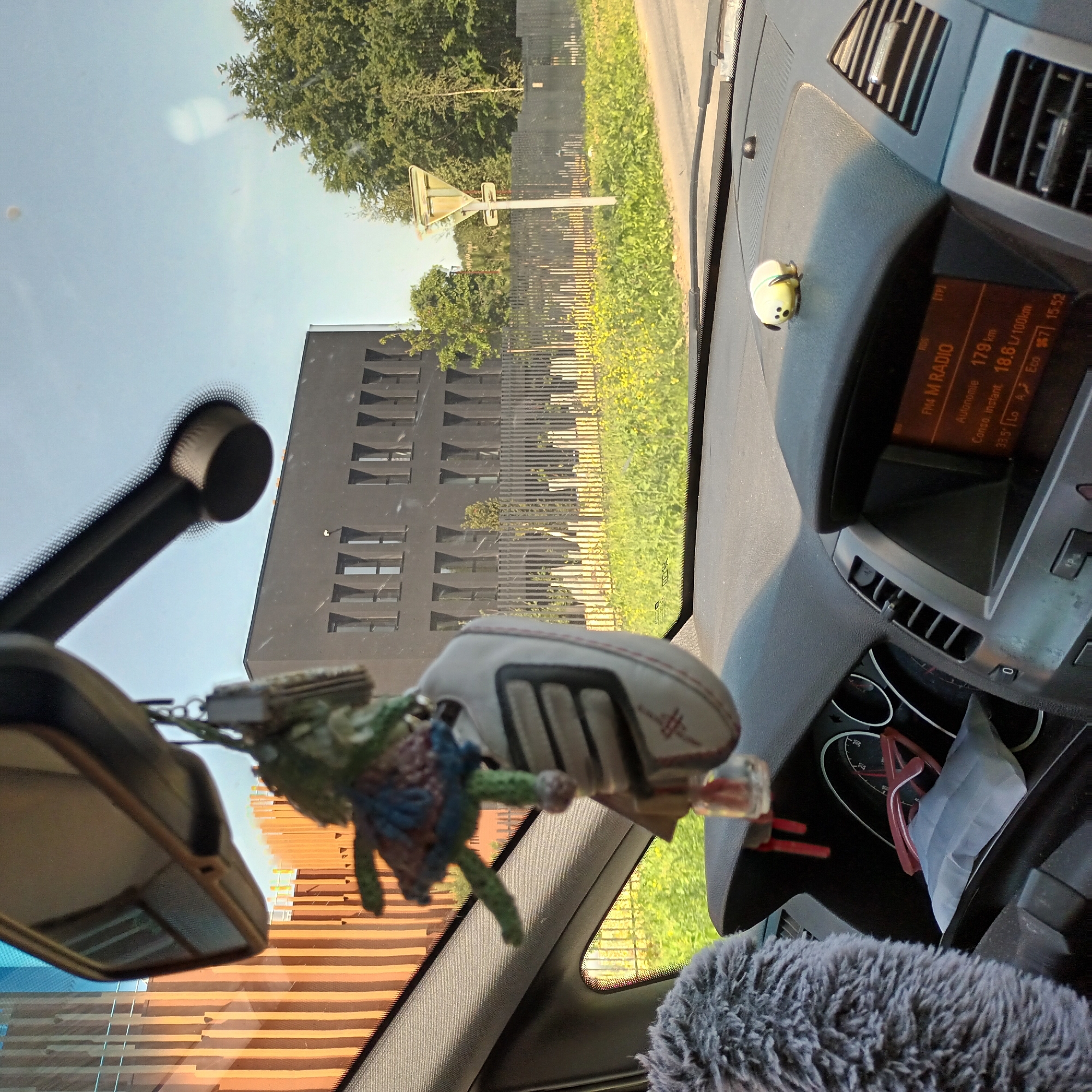The image is taken from the interior of a car, looking out the windshield. A rearview mirror and a fuzzy steering wheel cover are visible in the foreground. Hanging from the mirror is a small, crocheted item. Through the windshield, a modern, dark-colored building with a distinctive window pattern is visible. There is also a street sign and some greenery outside the car. The car's dashboard is in view, including a display showing "FM4 M RADIO" and "179 km". The general environment appears to be urban or suburban, with a mix of buildings and vegetation.