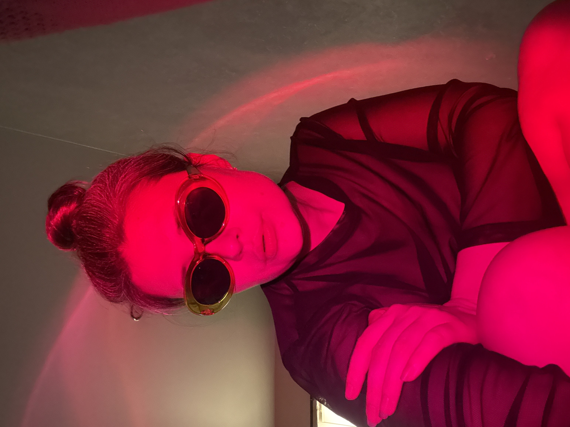 An indoor portrait captures a person, likely female, posing under intense red lighting. The subject is seen from the chest up, with their head tilted, looking towards the viewer. They wear round sunglasses with golden frames and dark lenses, obscuring their eyes. Their dark hair is pulled back into a bun or ponytail. The person is dressed in a sheer black top or dress, with elements of black undergarments or straps visible underneath. Their lips are slightly parted, and their expression is largely neutral, conveying a stylized or contemplative mood.
The entire scene is bathed in a strong, vibrant red light, which casts deep crimson hues over the subject's skin, hair, and clothing, creating dramatic shadows and highlights. This artificial illumination creates an atmospheric and mysterious ambiance. In the background, parts of a plain wall or ceiling are visible, also illuminated by the red light. Subtle arc-like patterns of red light can be seen on the upper wall, indicating a specific light source or projection. One of the subject's hands is visible, resting on their body, also intensely red from the lighting. The time of day is indiscernible due to the artificial light, but it appears to be a darkened room. There are no specific location cues within the image to suggest the provided context of Chaikovsky, Russia. No text is visible in the media file.