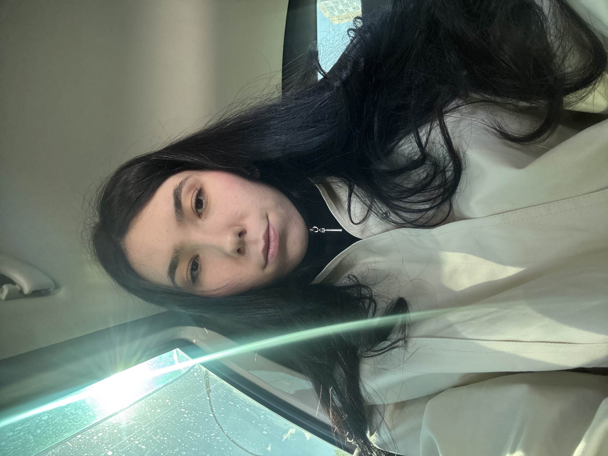 A young woman with long, dark hair is seated in a car. She is wearing a light-colored coat over a black turtleneck sweater. Sunlight is reflecting off the car's windshield. The location is not evident. There are no interactions visible.