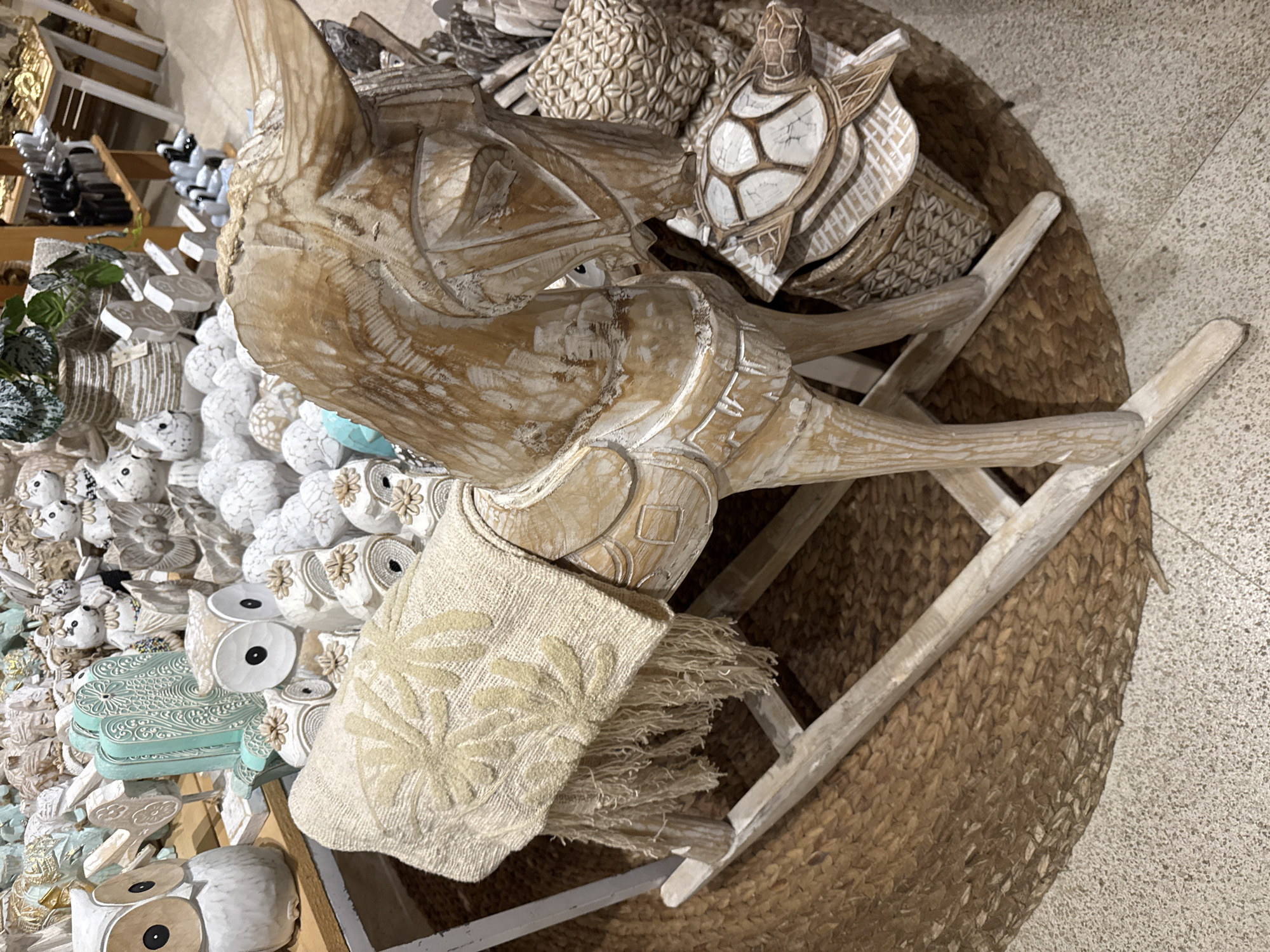 The scene is an indoor retail setting, potentially in Unknown, Indonesia, showcasing a variety of decorative items. Dominating the foreground is a large, carved wooden figure of a rhinoceros. Adjacent to the rhino is a white wooden turtle. The setting includes a woven basket serving as a base and a small white ladder. The left side of the image features a display of decorative owl figures, some in white, others in turquoise. The lighting is diffused and even. The scene conveys a commercial environment, possibly a store or market, with a focus on handcrafted and decorative goods.