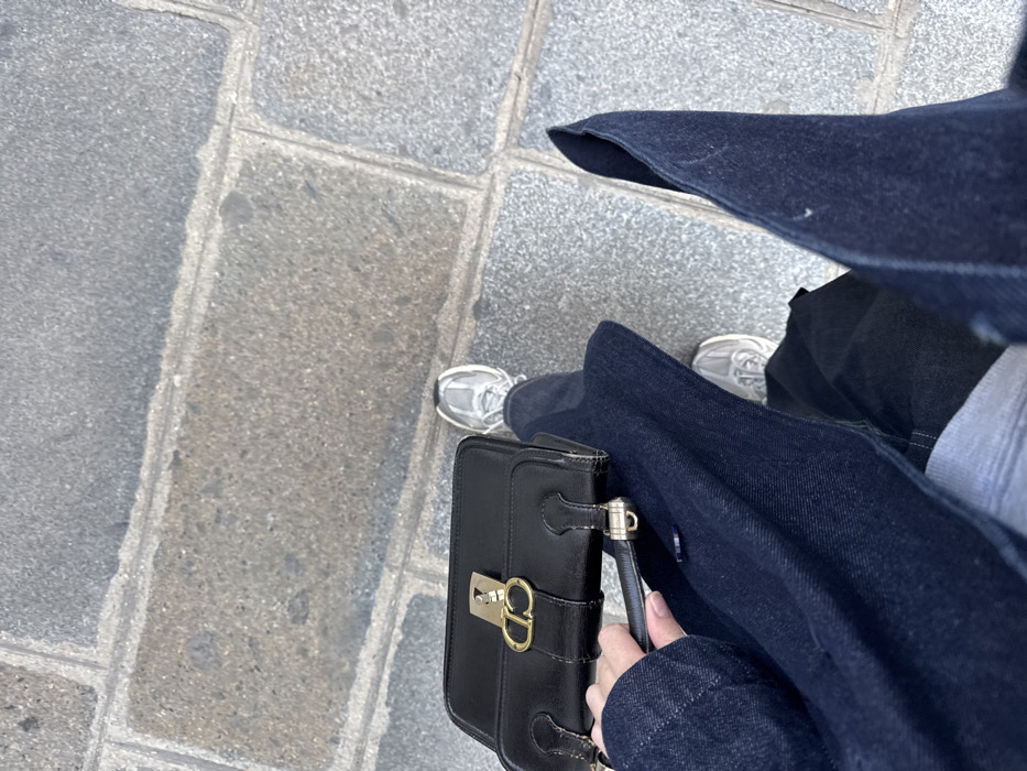 The image provides a high-angle, top-down view of an individual's lower body and right arm. The person wears dark blue denim outerwear, dark blue denim trousers, and light grey athletic shoes on both feet. A light grey undergarment is visible between the denim layers. The right hand holds a black leather handbag featuring a gold-toned metal clasp designed with an interlocking "CD" logo, characteristic of the Christian Dior brand. The ground is paved with grey, rectangular stone blocks, a common architectural element of sidewalks and public spaces throughout Paris, France. The precise location within the city of Paris cannot be determined from the visible elements.