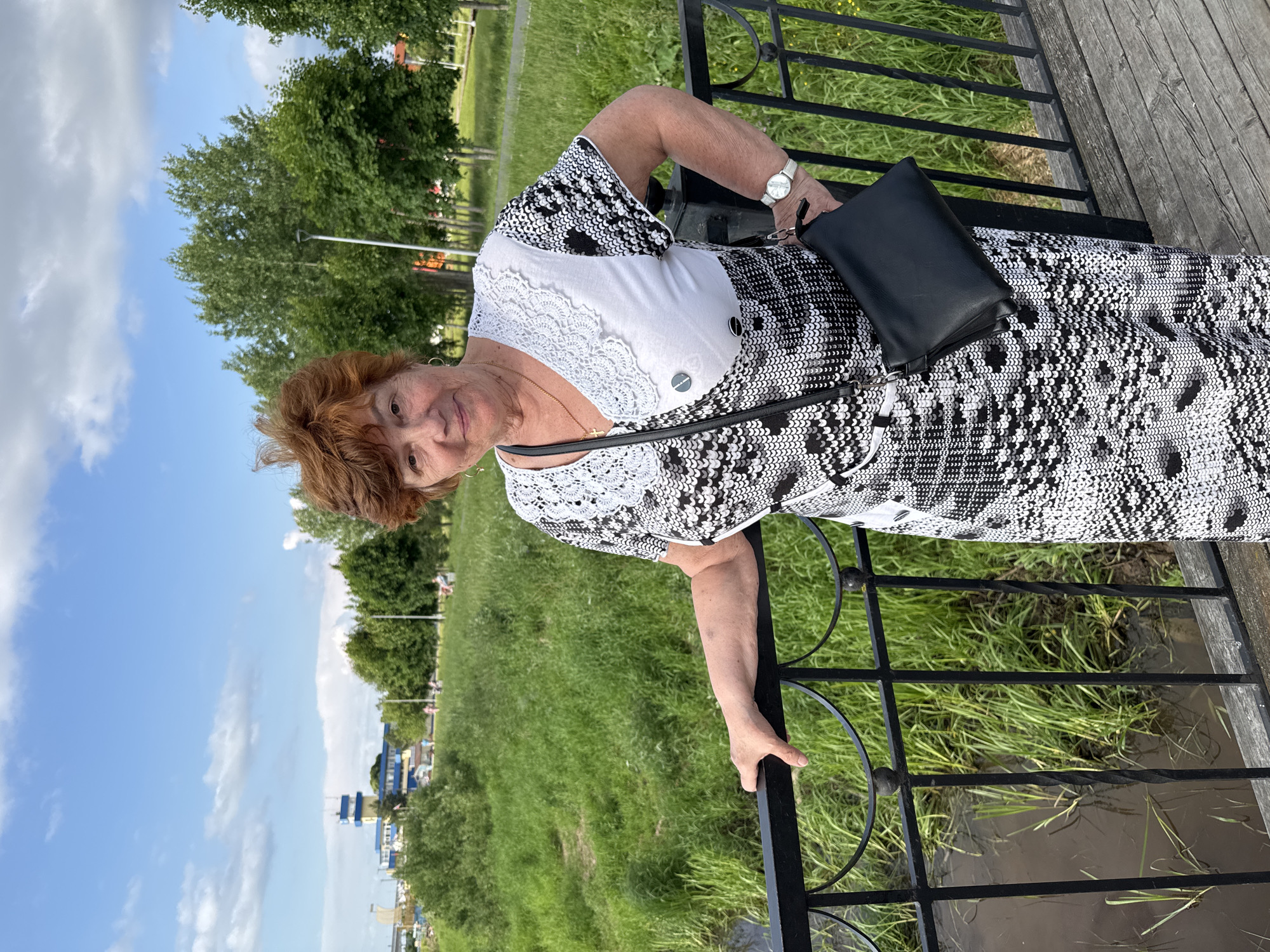 A woman with short, reddish-brown hair and a fair complexion stands on a wooden bridge with a black metal railing. She is wearing a black and white patterned dress and a black purse slung over her shoulder. Her left hand rests on the railing, and her right arm is bent, with her hand near her side. She is smiling and looking at the camera. Behind her, a grassy embankment slopes down to a dark body of water. In the background, there are trees, a lamppost, and a blue and yellow building with a tall tower. The sky is blue with scattered white clouds. The image appears to have been taken in an outdoor park setting in the city of Kirishi, Russia.