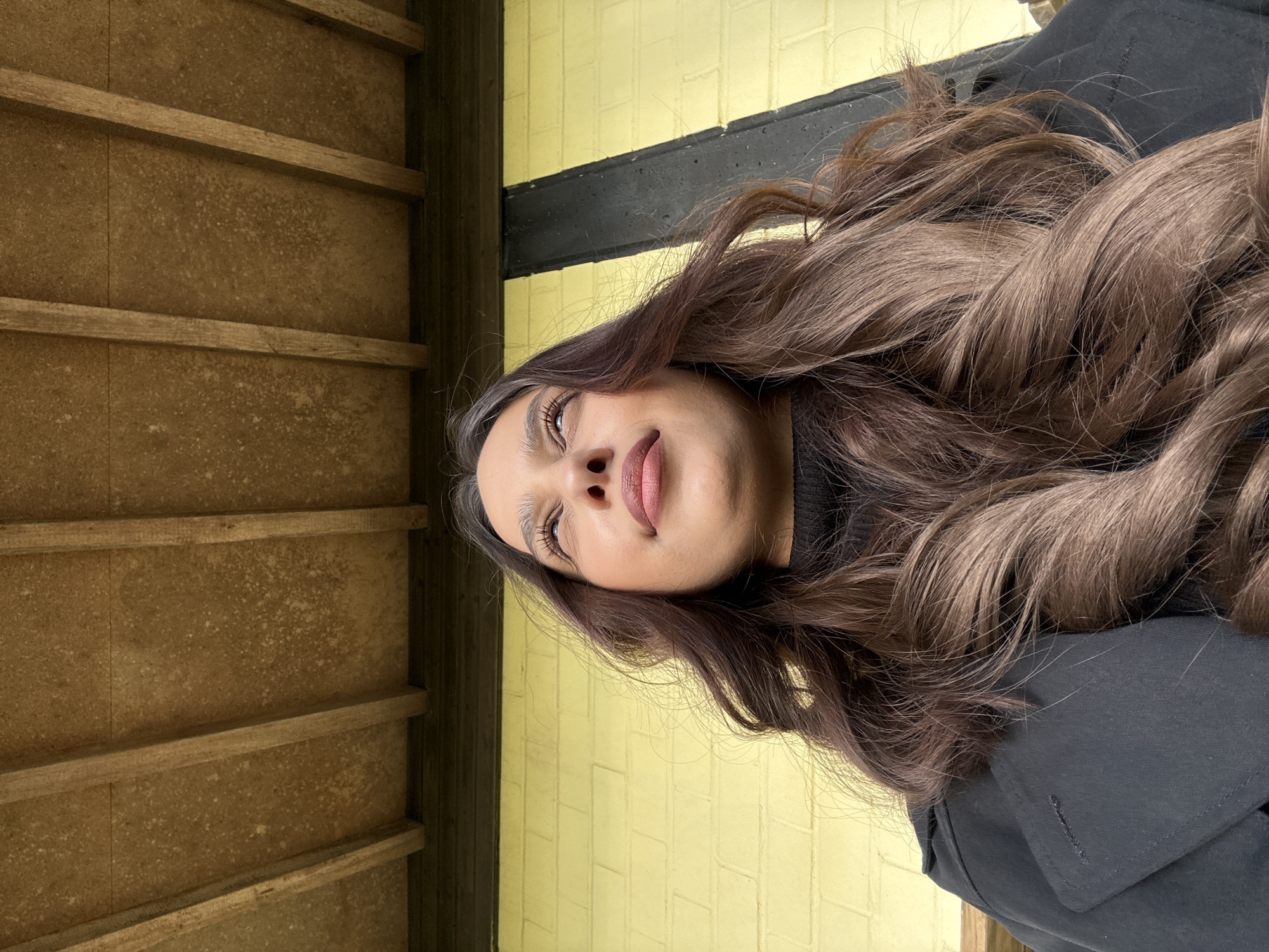 A young woman with long, brown wavy hair is pictured in an outdoor setting. She wears a black top and a black coat. The background consists of a yellow brick wall with a black stripe, and a section of brown wooden slats. The location is Wegberg, Germany.