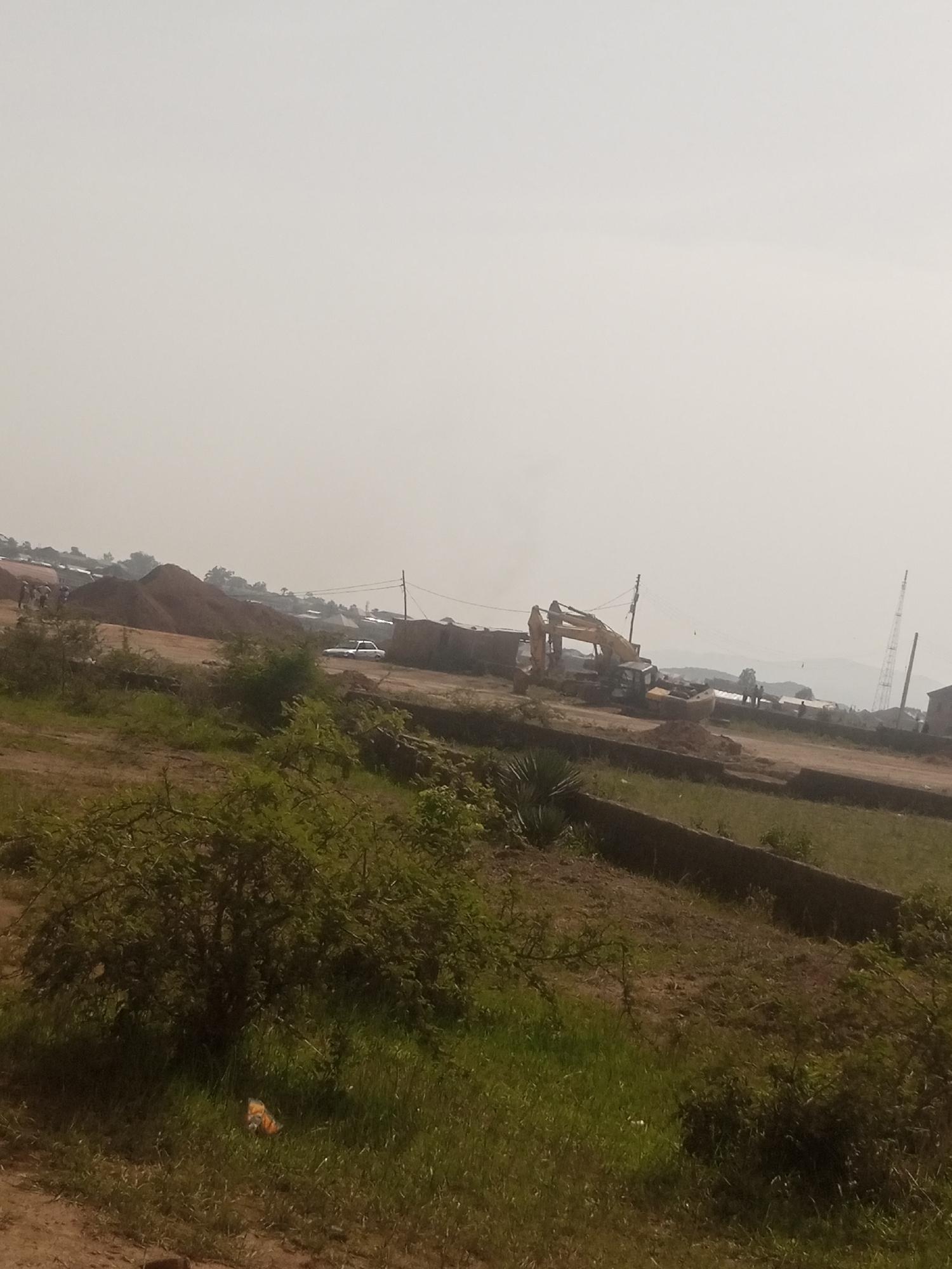 The image depicts a construction site in Jos, Nigeria, under a hazy sky. In the background, several structures and large mounds of earth are visible, suggesting ongoing development. A white car is parked on a dirt road. In the mid-ground, a yellow excavator is positioned near a pile of dirt, and other construction equipment is present. A few people are scattered around the site, appearing to be involved in the construction activities. In the foreground, there is dense green vegetation with a small, orange and black object partially obscured by the foliage. A low concrete wall runs horizontally across the scene, separating the foreground from the construction area. The overall impression is of an active construction zone in a somewhat rural or semi-urban setting.