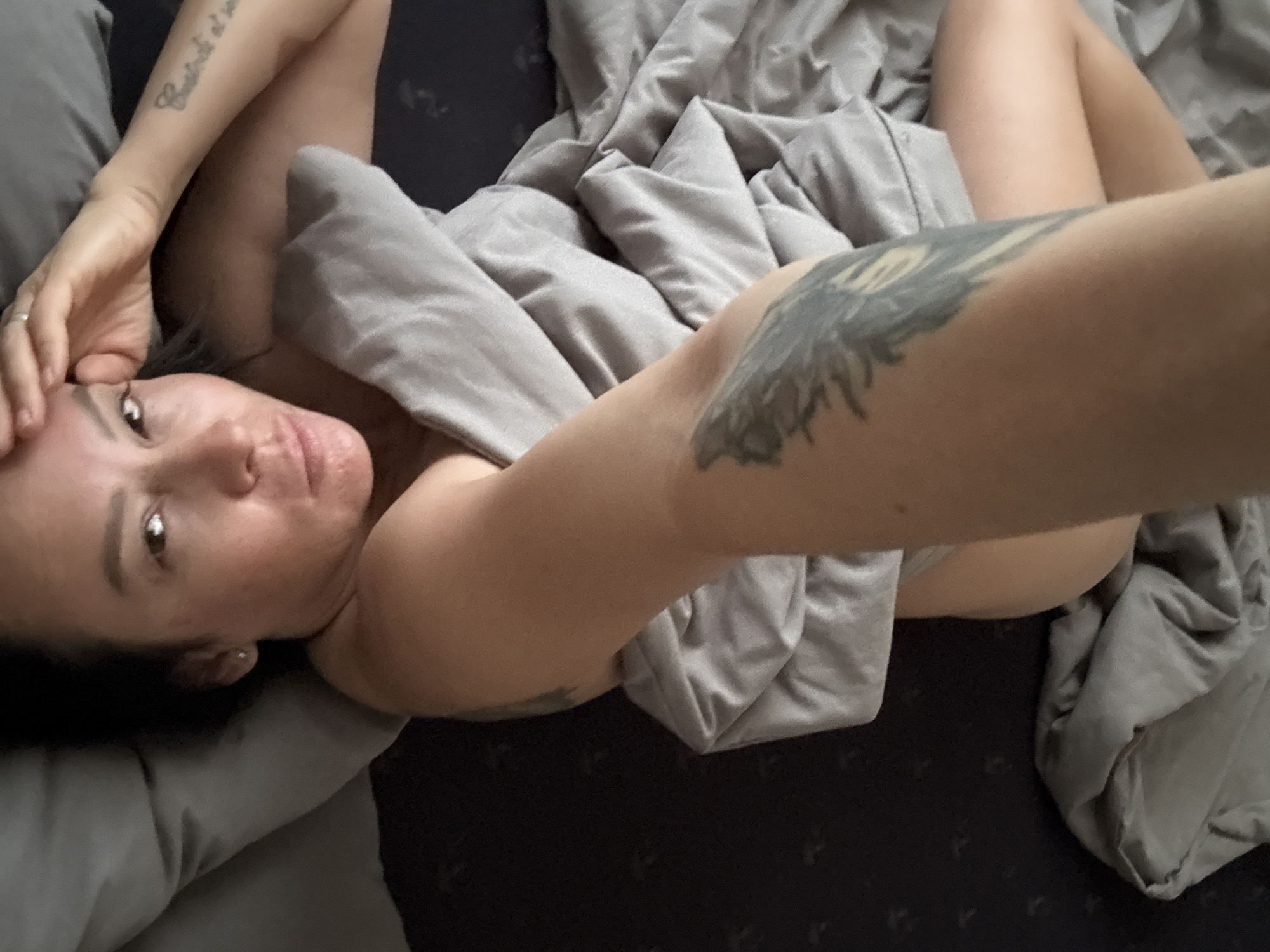 A woman with tattoos is lying in bed, partially covered by a gray blanket. She is resting her hand on her forehead. The location is not discernible in this image, and there is no evidence that it is taken in the city of Chaikovsky, Russia.