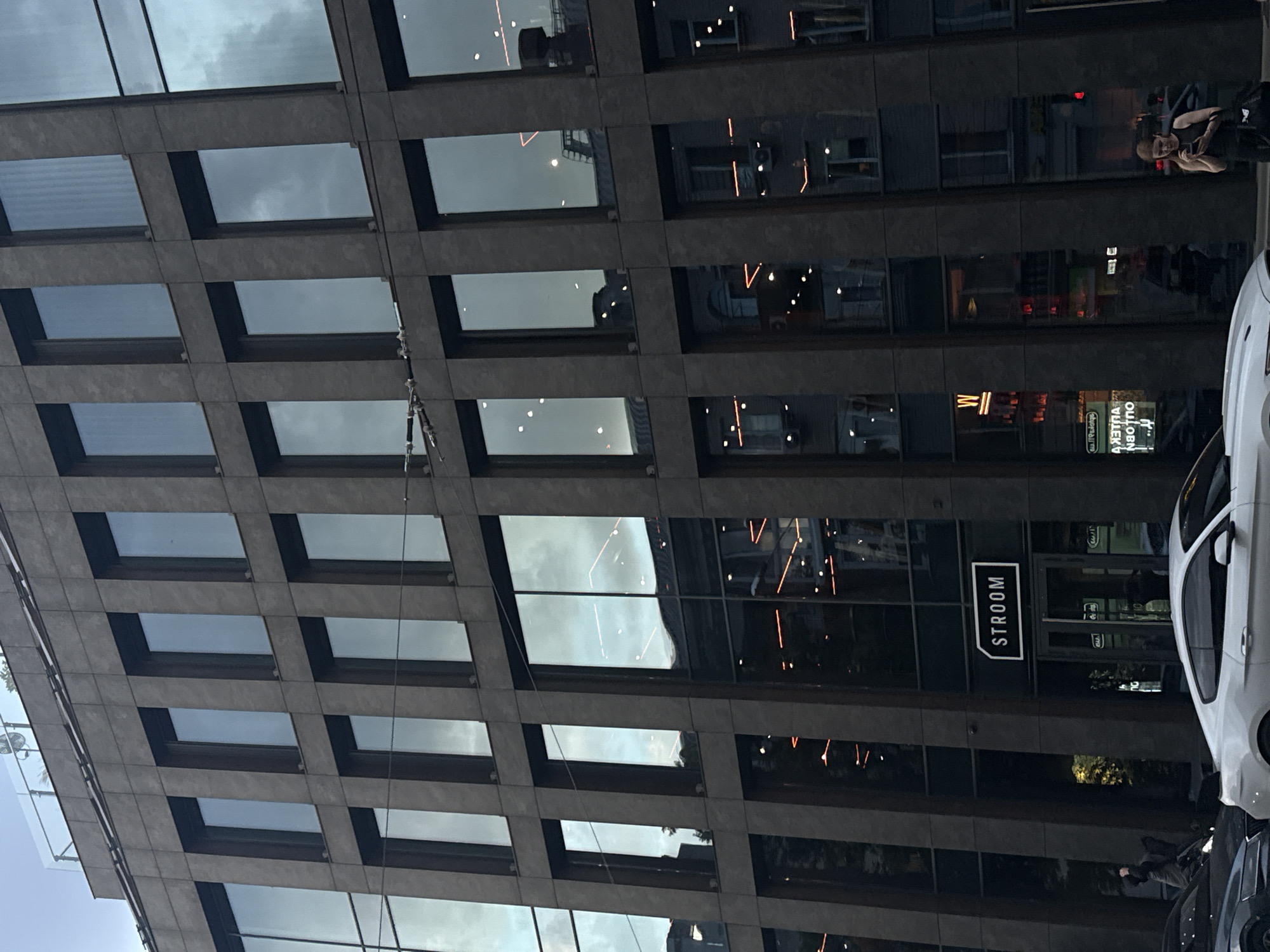 This is a ground-level, medium shot image of the facade of a modern commercial building. The building has a dark, textured exterior with a grid of rectangular windows. The windows reflect the sky, which appears overcast with some lighter patches. Some windows reveal interior lighting, including horizontal orange lights and circular white lights. A black sign with white lettering that reads "STROOM" is affixed above an entrance. Several cars are parked in front of the building, including a white sports car in the foreground. A woman is standing on the sidewalk to the right of the entrance, looking at her phone. Another person is partially visible on the sidewalk to the left, near a dark car. The setting is a street in Dnipro, Ukraine.