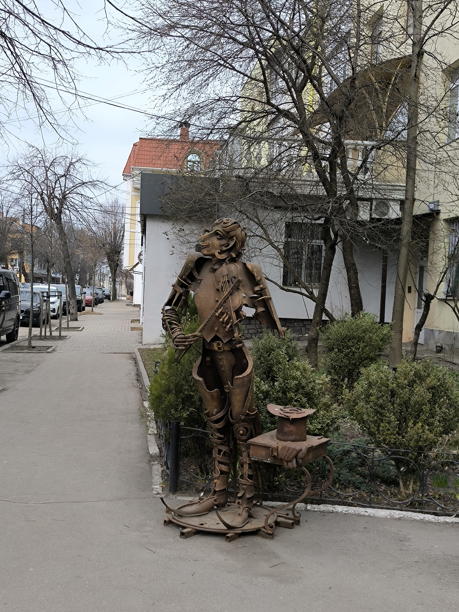 The image shows a metal statue of a man holding a violin, standing on the sidewalk in Vinnytsia, Ukraine. The statue is rusted and appears to be made of recycled metal parts. The statue is standing next to a small wrought iron table with a hat on top. Behind the statue are bushes and a building. The sidewalk is gray, and there are trees lining the street in the background. Several cars are parked along the side of the road. The sky is overcast.