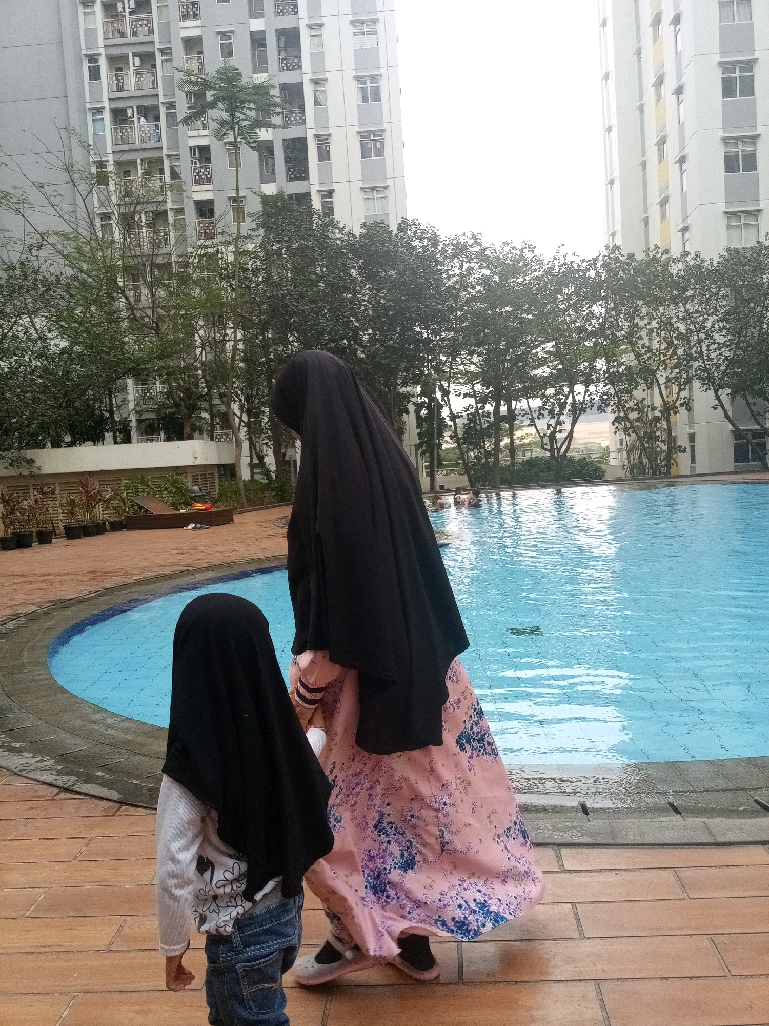 In Bekasi, Indonesia, a mother and child stand on a wooden deck next to an outdoor swimming pool. The mother, wearing a black hijab and a pink floral dress, stands to the right. To her left and slightly in front, a young child, also wearing a black hijab and blue jeans, faces away from the camera. In the background, two apartment buildings rise behind a line of trees. Some individuals can be seen swimming in the bright blue water of the pool. The deck appears wet, suggesting recent use of the pool.
