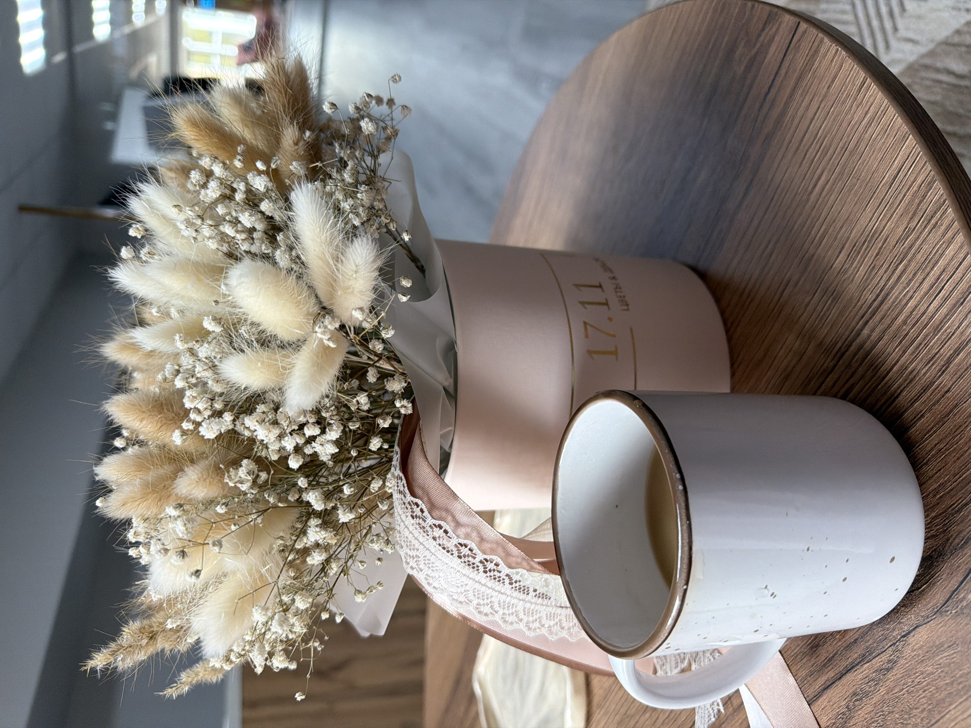 This indoor still life image, captured in Chaikovsky, Russia, showcases a dried flower bouquet and a mug on a round wooden table. The prominent bouquet features fluffy, light brown and cream-colored lagurus ovatus (bunny tail grass) mixed with delicate white gypsophila (baby's breath) and some spiky dried grasses. It is arranged in a light pink cylindrical box, tied with light pink satin and white lace ribbons that cascade onto the table. Gold text on the box clearly displays "17.11", followed by "ЦВЕТЫ И &" (FLOWERS AND &), with further text unreadable. Adjacent to the flower box, a white enamel mug with a brown rim and small dark speckles sits on the table; it appears empty. The table is a light brown wooden surface with a visible grain. The background is blurred, showing bright, out-of-focus light, indicating a daytime interior. No people or specific activities are depicted, presenting a tranquil arrangement of objects.