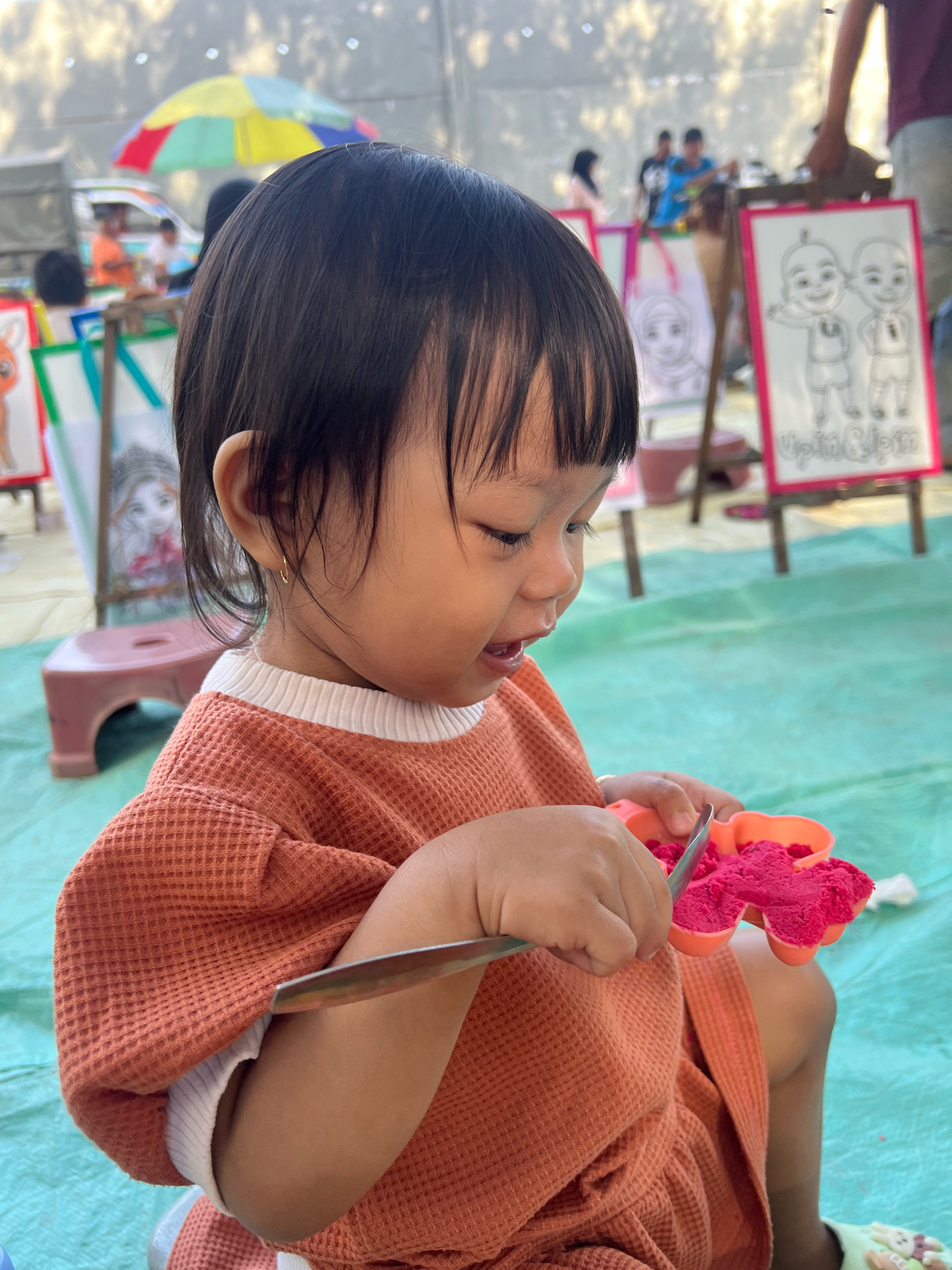 A young child, likely a girl, with dark hair and bangs, wears an orange waffle-knit shirt and is engrossed in playing with bright pink kinetic sand inside a heart-shaped mold. She holds a small silver tool, possibly a spatula or spoon, and appears to be scooping or manipulating the sand. The child is seated outdoors on a teal-colored tarp. In the background, several easels are set up with drawings, including cartoons of characters that resemble "Upin & Ipin." Other children and adults are visible in the distance, with a colorful umbrella providing shade over a vehicle. The setting suggests an outdoor art or craft activity, possibly a community event or marketplace, in Kecamatan Pati, Indonesia.