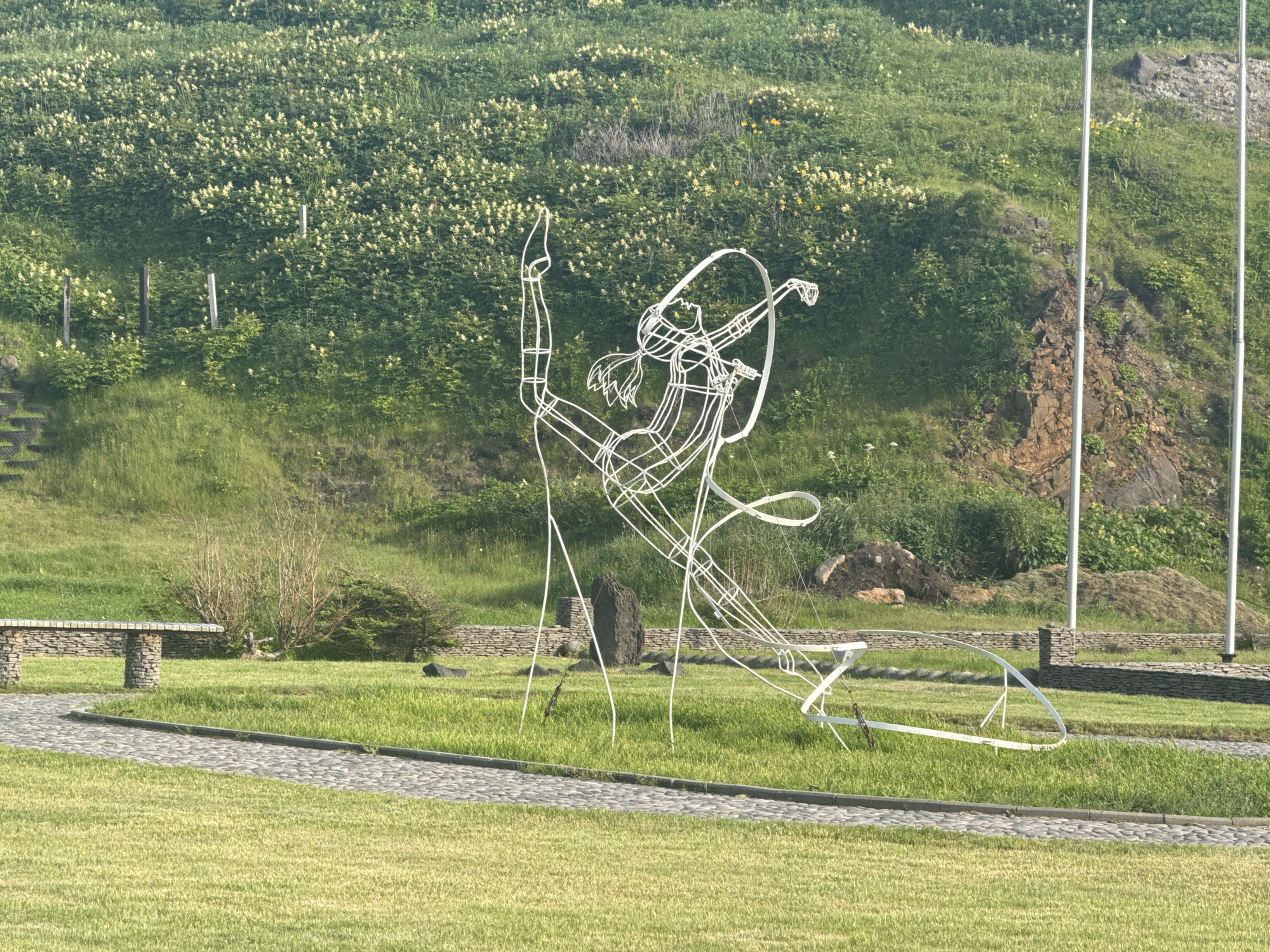A wire sculpture depicting a rhythmic gymnast in motion is positioned in a park in Kuril'sk, Russia. The sculpture is white and abstract, with the gymnast's body outlined in thin wire, and a large hoop is integrated into the design. The gymnast is in a dynamic pose, with one leg extended upwards and arms in a fluid motion, suggesting the performance of a routine. A large hoop, also made of wire, is held in one hand and curves around the gymnast.
The sculpture is situated on a well-maintained lawn, with a cobblestone path curving around it. To the left of the sculpture, a stone bench with a stone table rests on a paved area. In the background, a steep, grassy hillside is covered with lush green vegetation dotted with small white flowers. Several wooden posts and a dark, oblong stone are also visible in the immediate vicinity of the sculpture. Further up the hill, rocky outcrops and more vegetation are present. Two tall, slender poles stand vertically on the right side of the frame, their purpose unclear. The lighting suggests it is daytime, possibly morning or late afternoon, with soft sunlight illuminating the scene. The overall impression is one of an outdoor public space, likely a park or recreational area, featuring artistic elements integrated into the natural landscape.